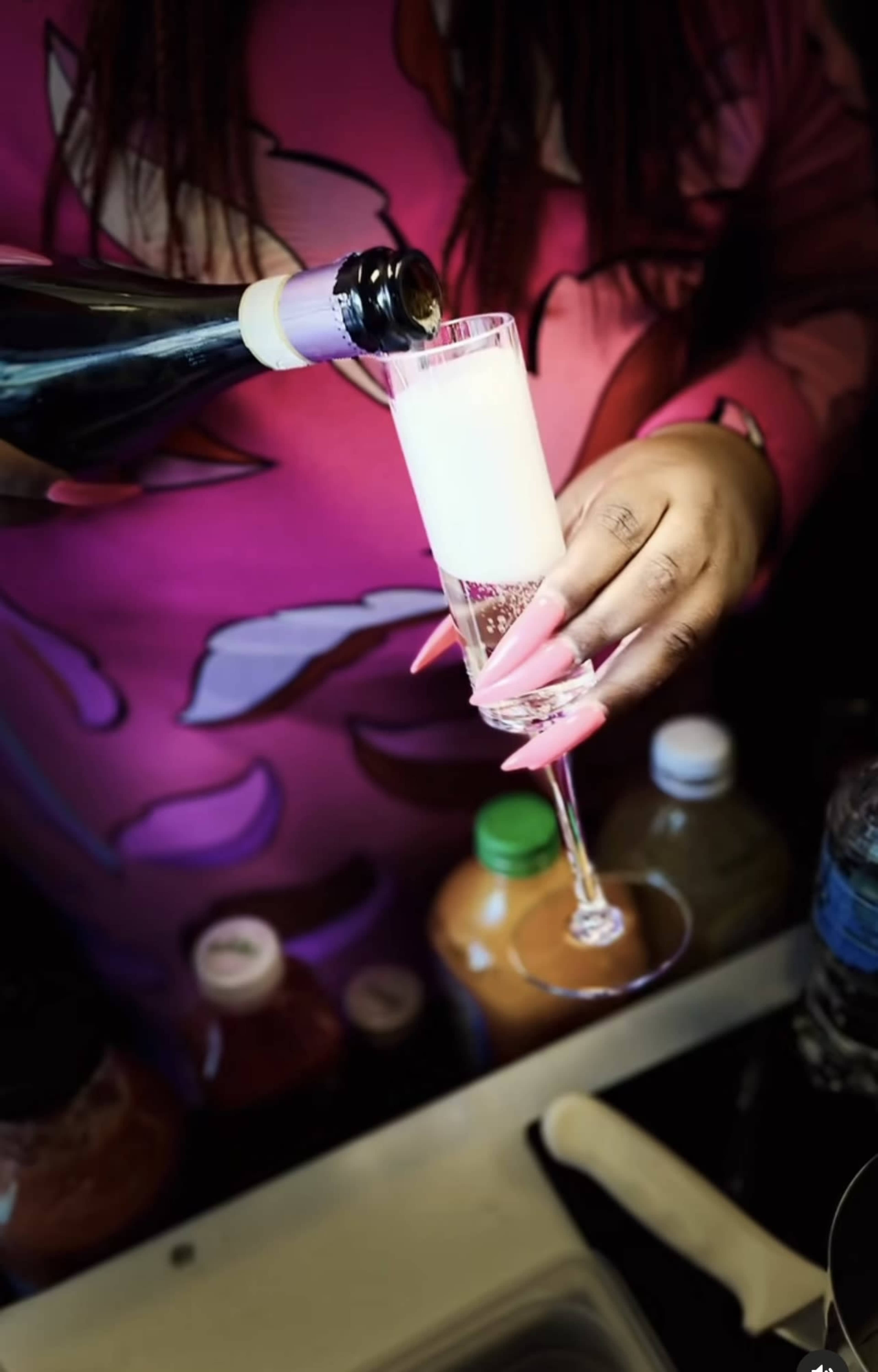 A person with long nails is pouring a sparkling beverage into a champagne flute, surrounded by various drinks in the background.