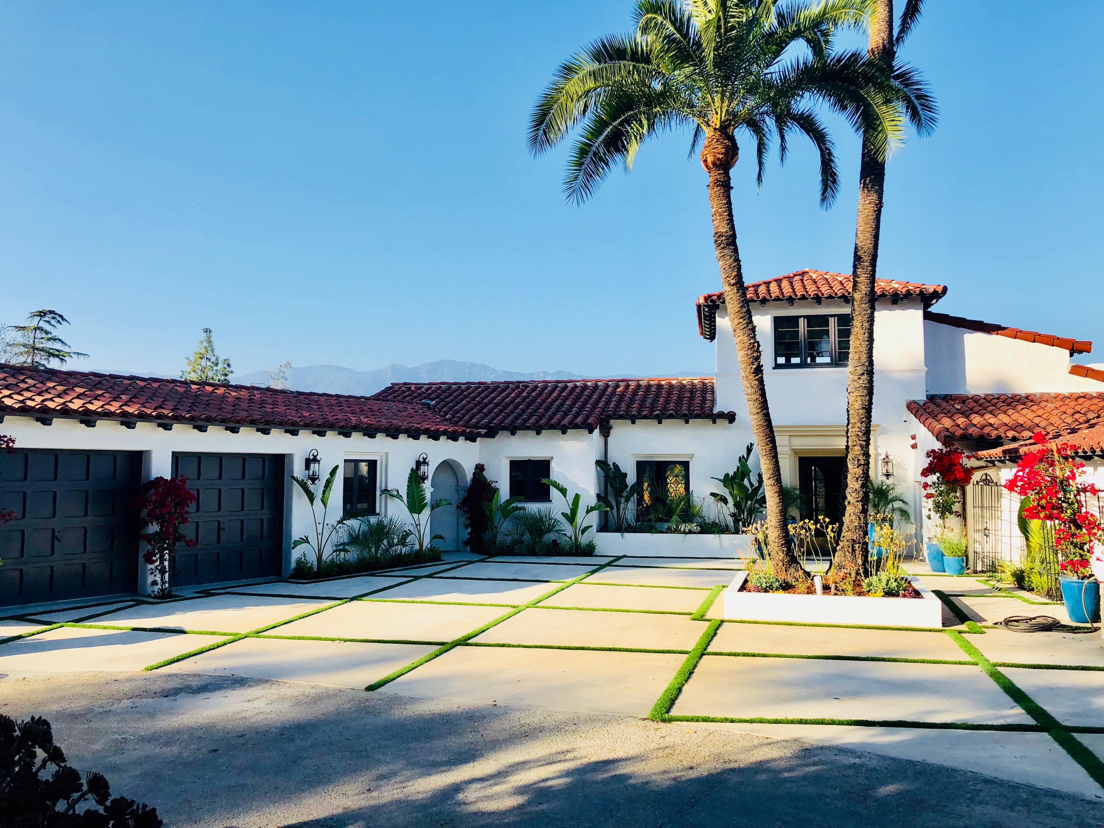 A Mediterranean-style house with a tile roof is set in a landscaped courtyard featuring palm trees and decorative plants.