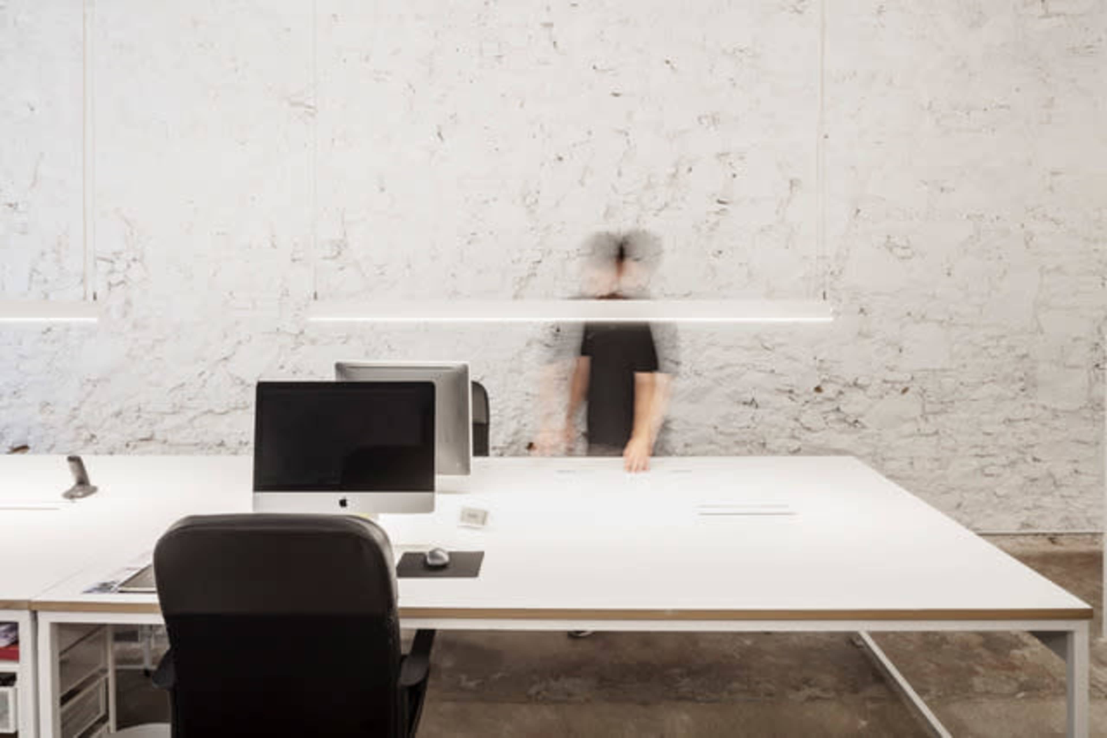 A person walks in a minimalist office space with a white desk and two desktop computers against a textured wall.