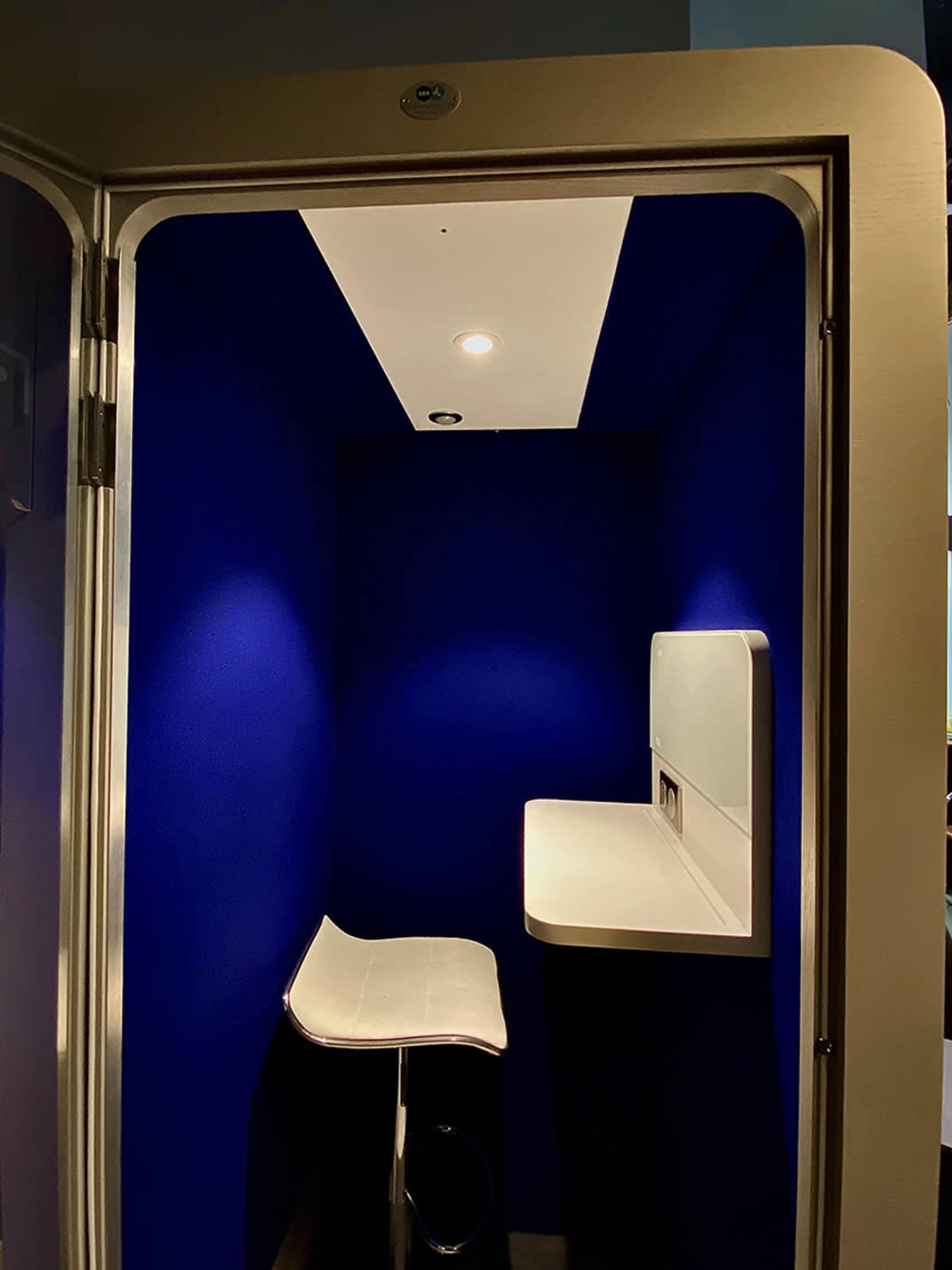 The image shows a small booth with a blue interior, featuring a white counter and a stool.