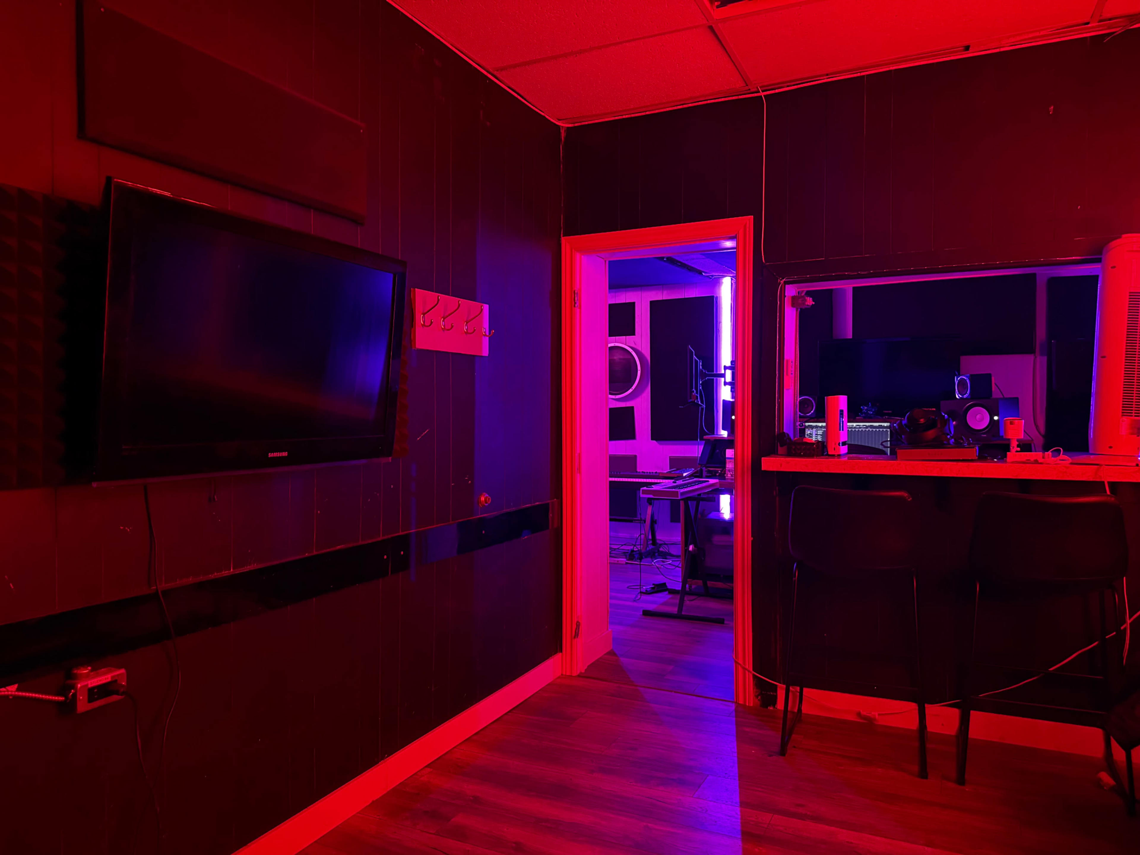 The image shows a dimly lit room with red and purple lighting, featuring a television on the wall, a bar area with stools, and a doorway leading to another room equipped for audio production.