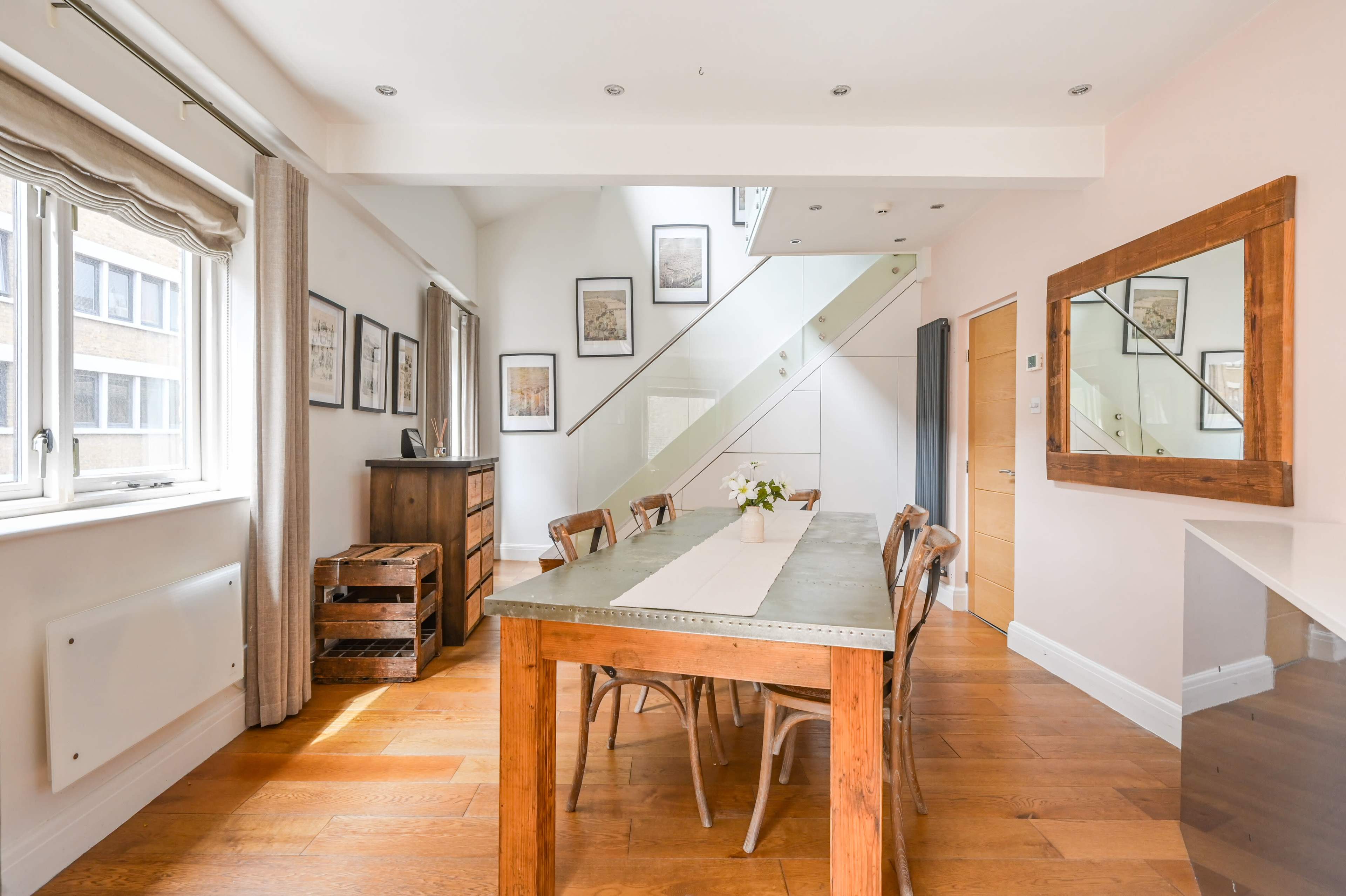 A modern dining area features a long wooden table surrounded by chairs, with large windows letting in natural light and framed artwork on the walls.
