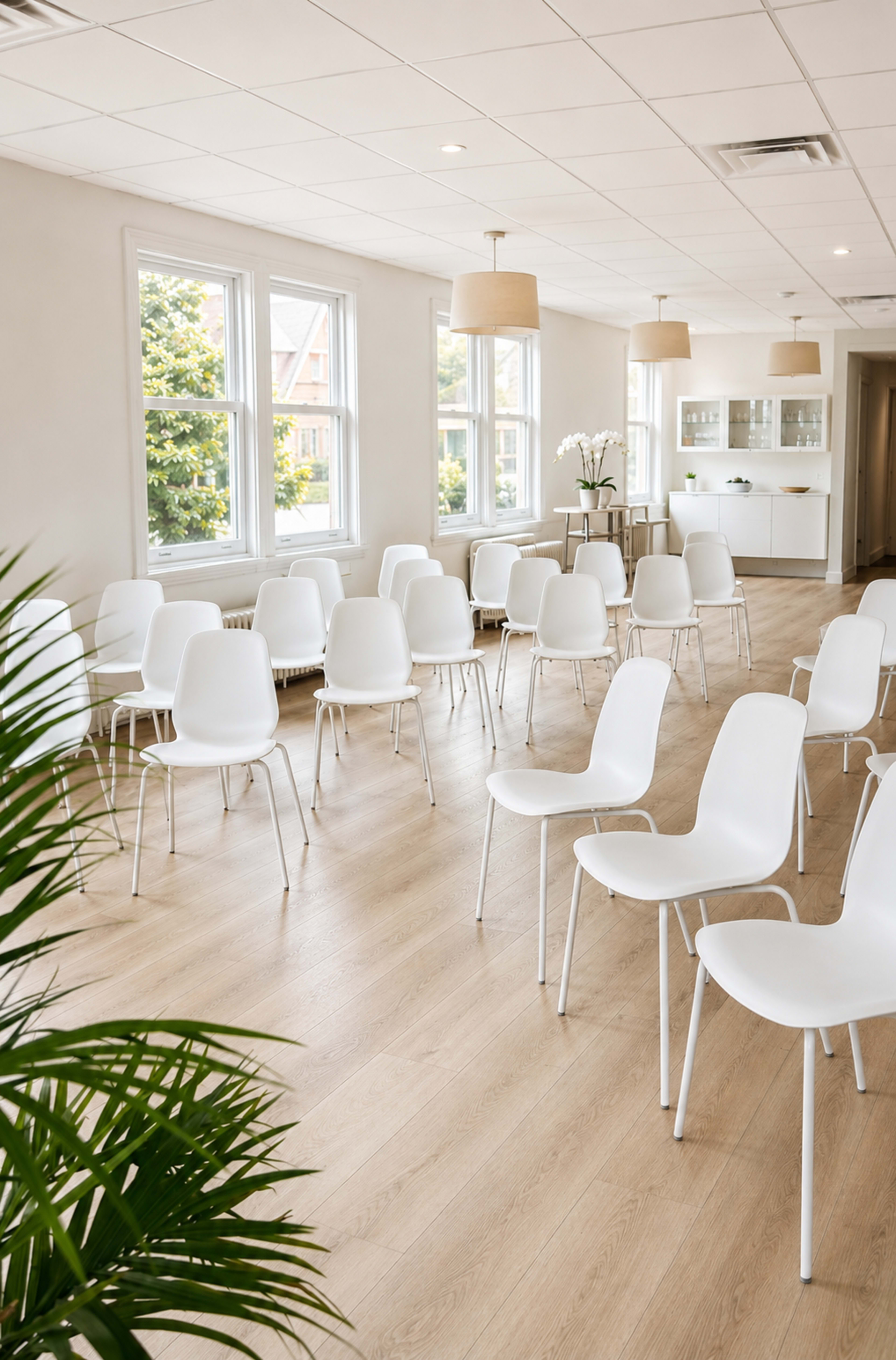 The image shows a minimalist room with several white chairs arranged in rows, large windows letting in natural light, and a small seating area in the background.