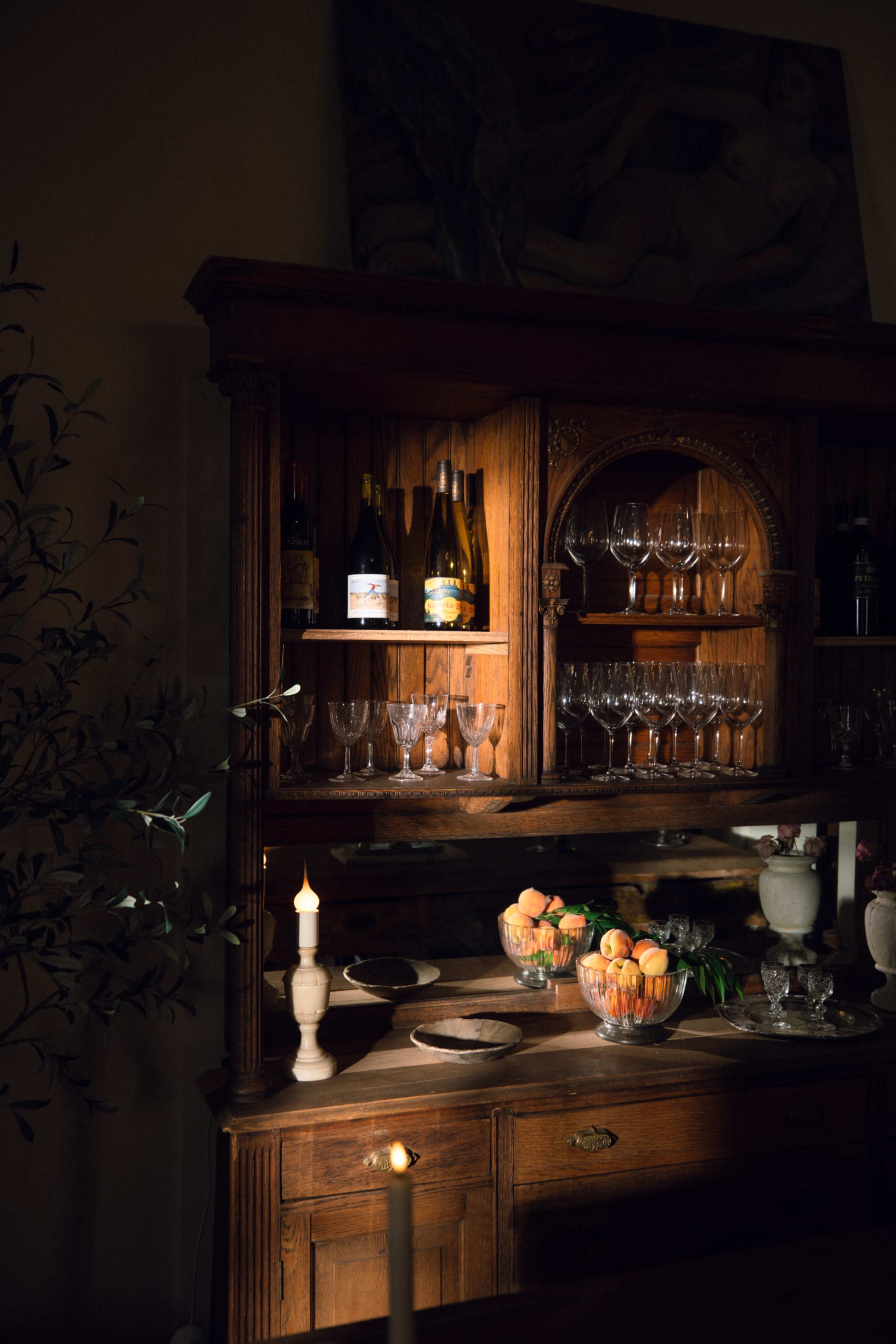 A wooden glass cabinet displays bottles of wine, wine glasses, and a bowl of peaches, illuminated by soft lighting and a candle.