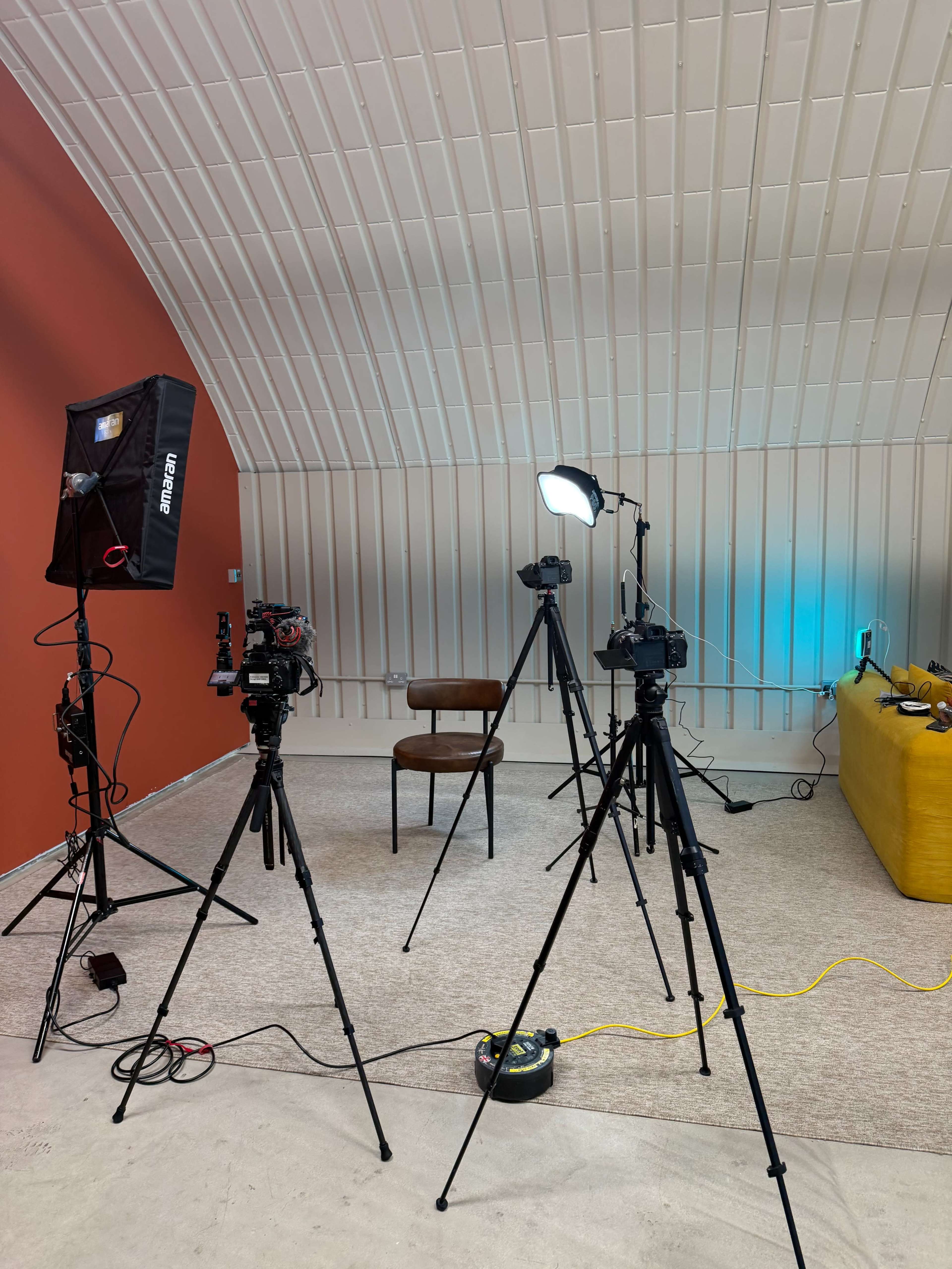 The image shows a studio setup with three cameras on tripods, a chair in the center, and various lighting equipment against a backdrop of red and white walls.