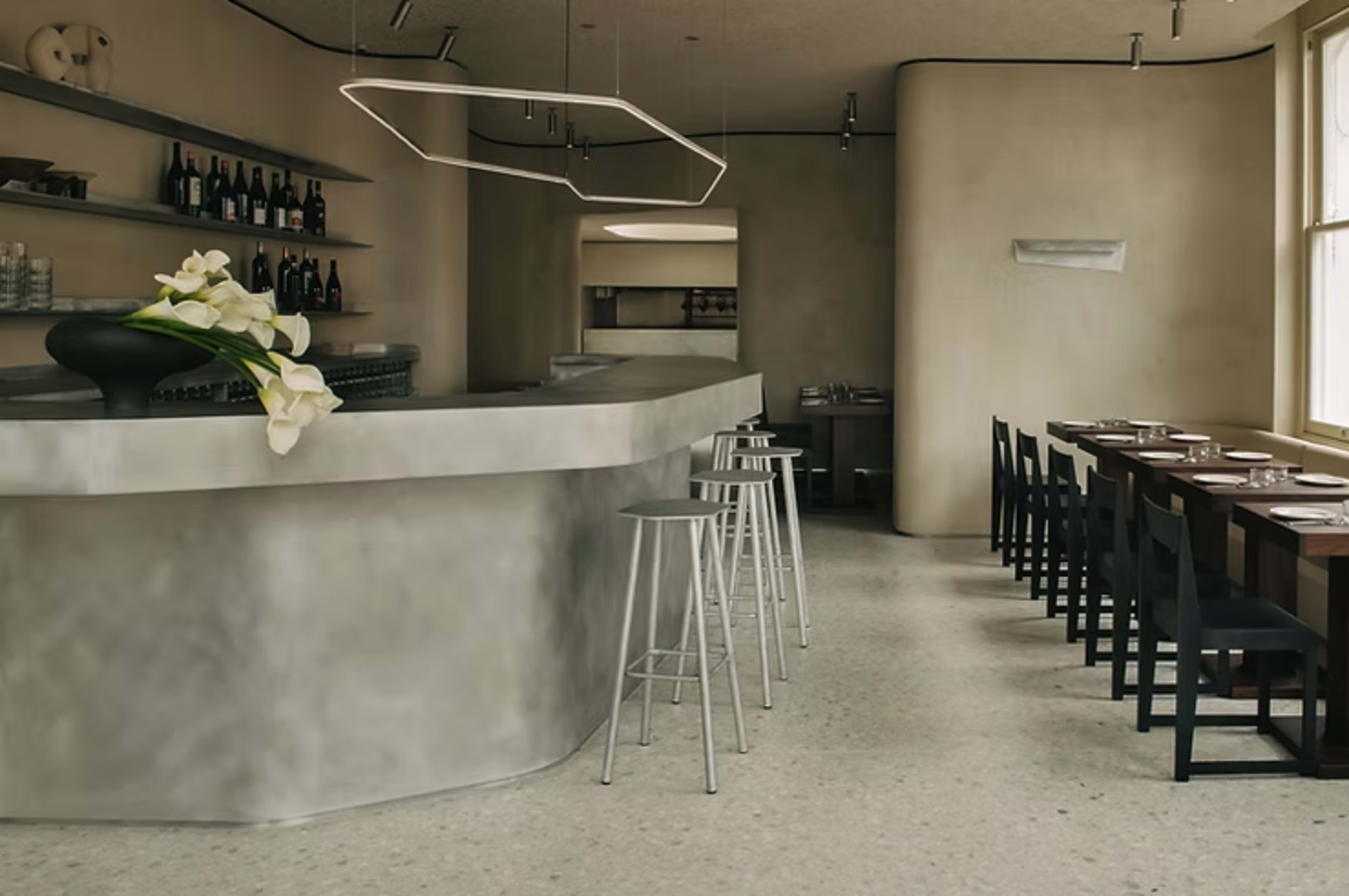 The image shows a modern, minimalist restaurant interior featuring a curved bar, tall stools, and a row of tables along the wall.