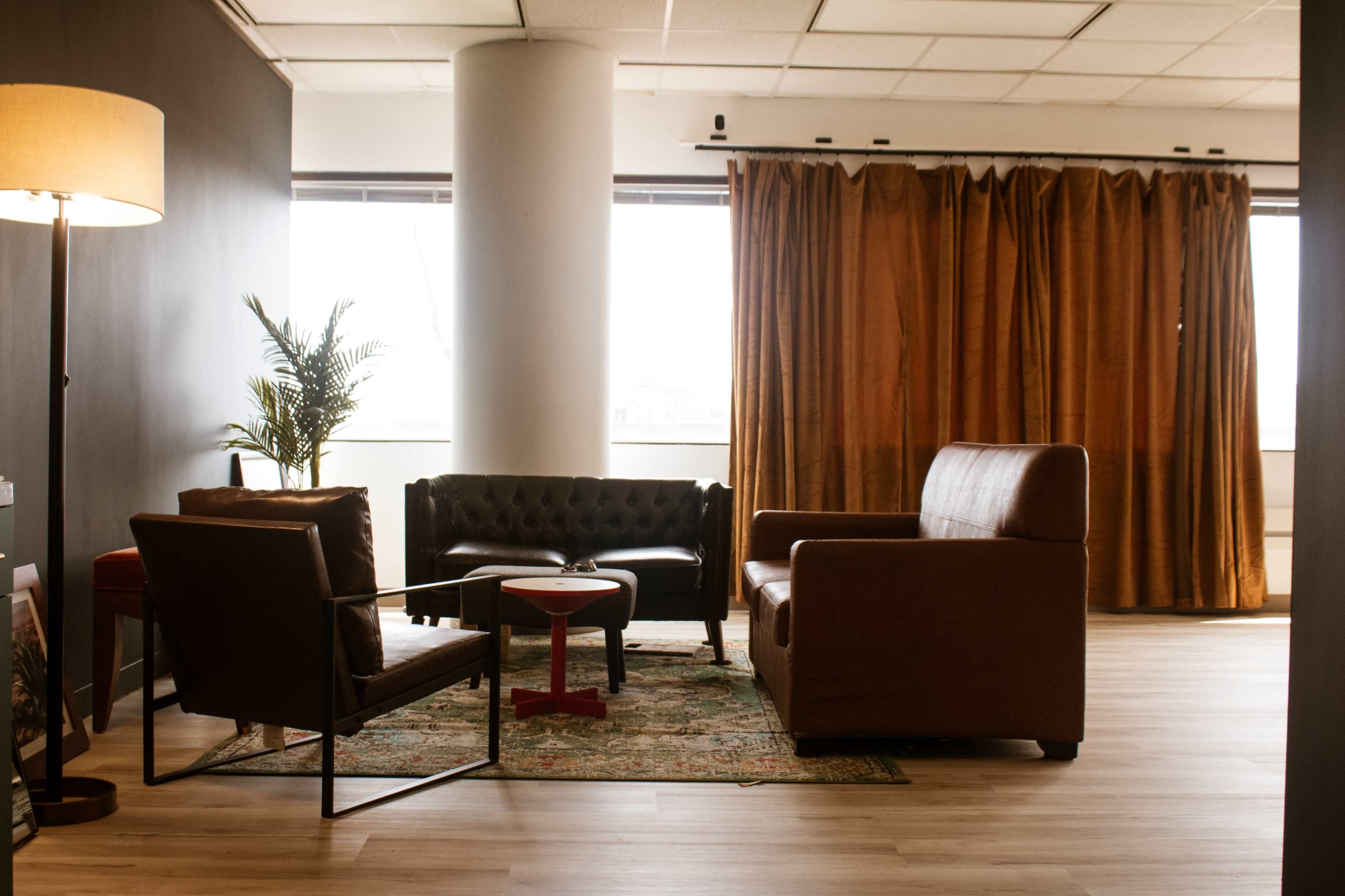 The image shows a cozy lounge area with two chairs, a sofa, and a small red table, set against a backdrop of large windows and brown curtains.