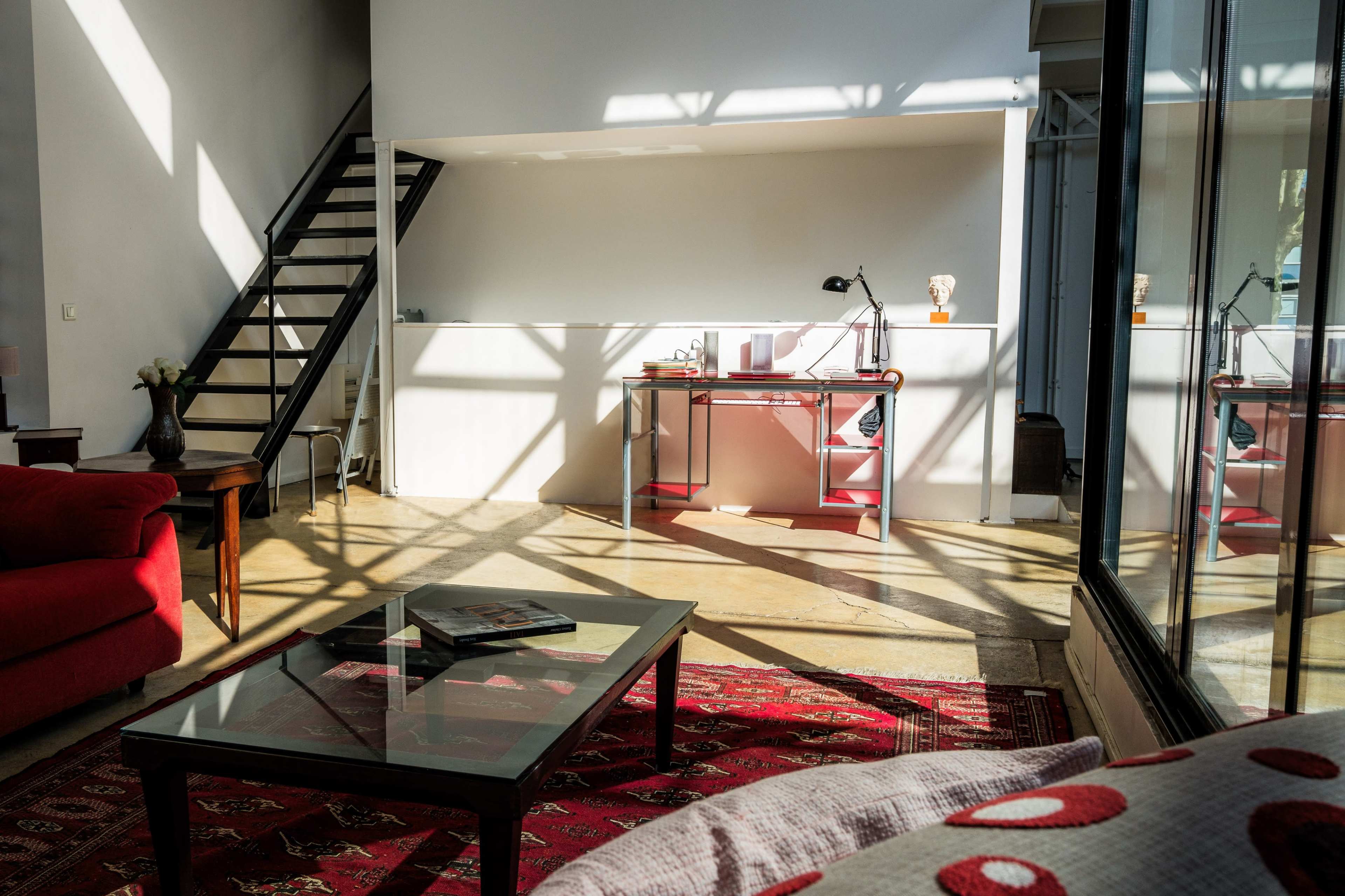 A sunlit living area features a red sofa, a glass coffee table, and a workstation set against a backdrop of shadows cast by nearby stairs.
