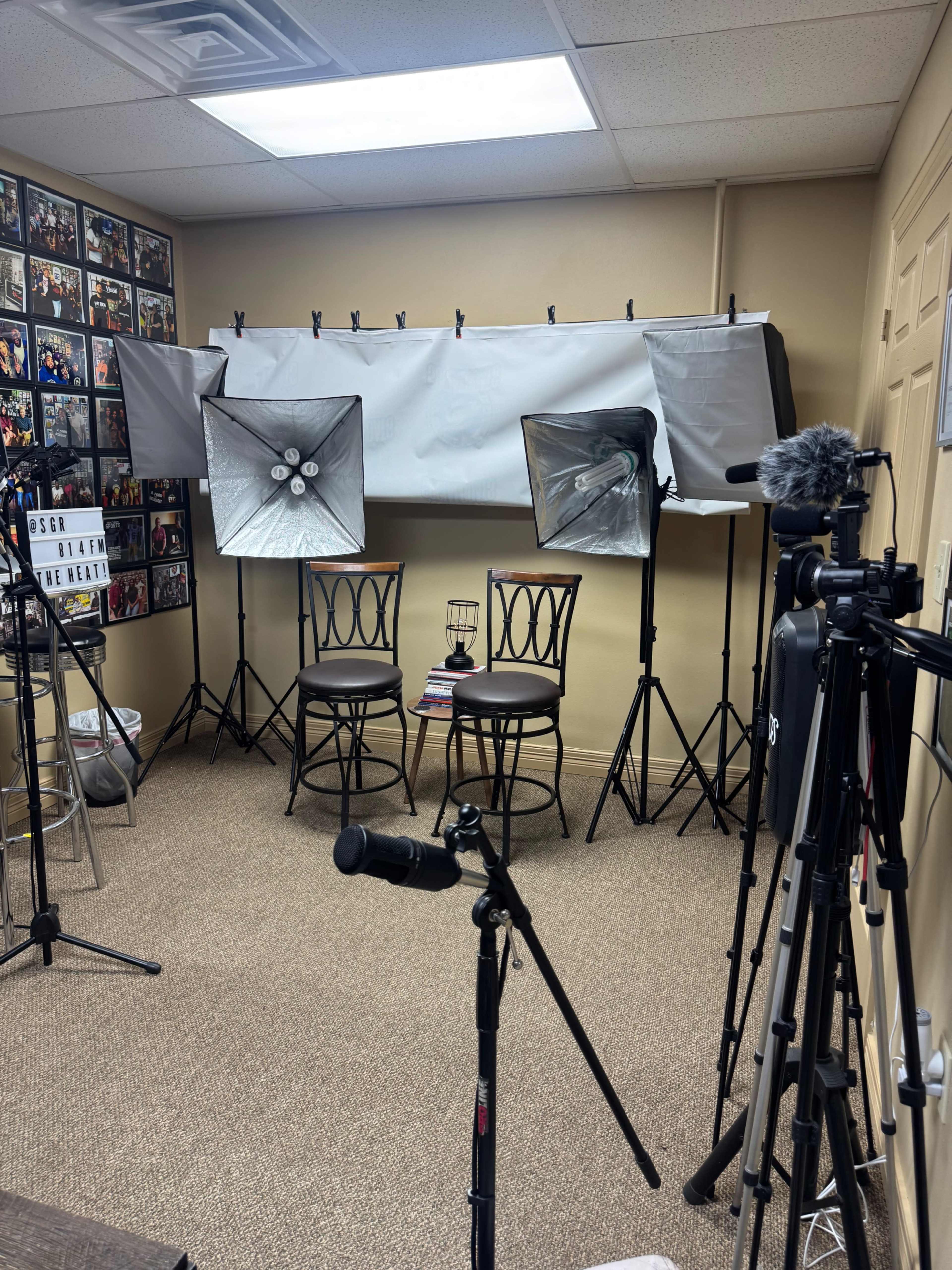 The image shows a small studio setup with two chairs, lighting equipment, and camera tripods arranged against a wall filled with framed pictures.