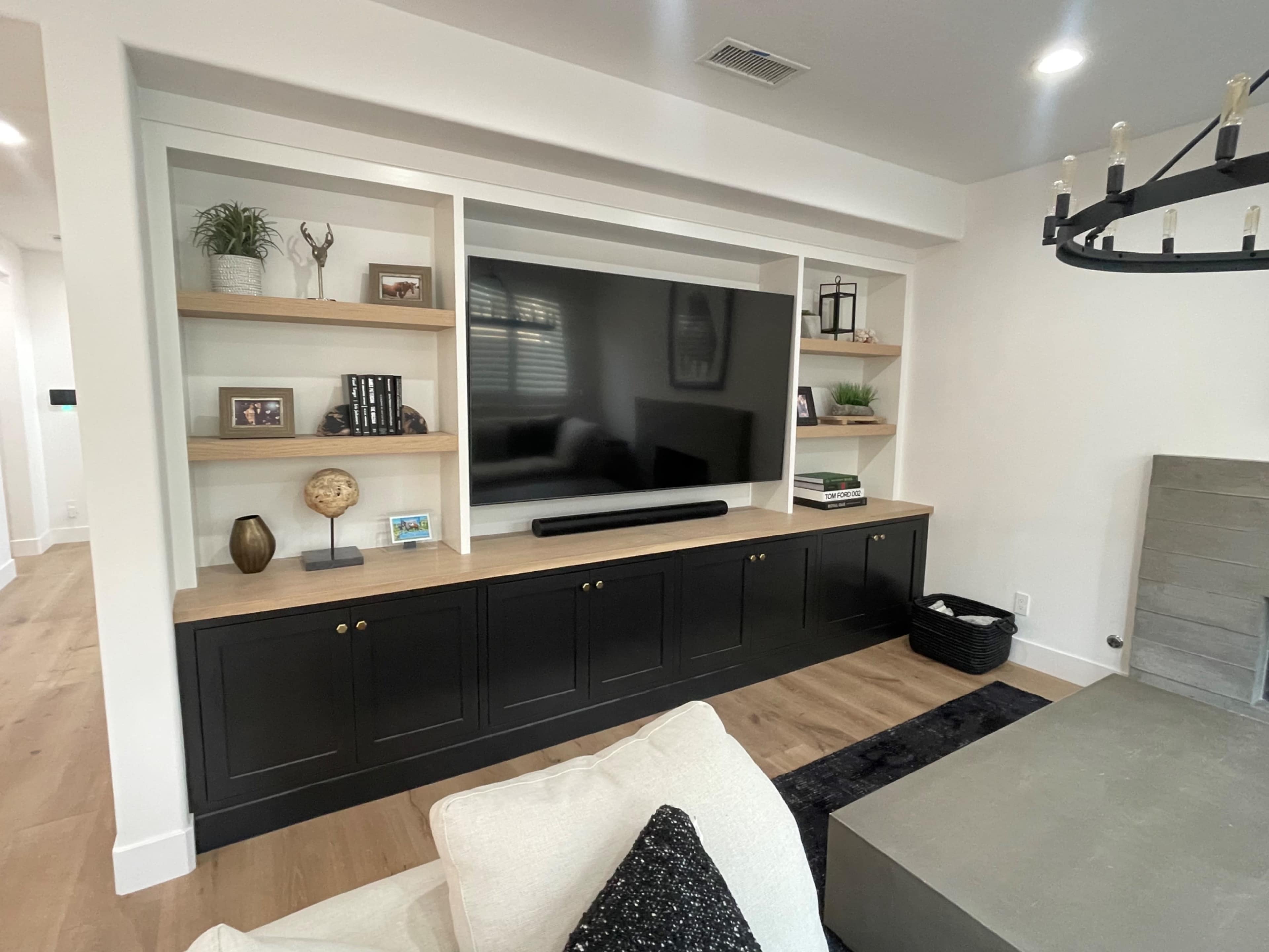 A modern living room with a large wall-mounted TV integrated into a custom wooden entertainment center featuring open shelving and a black cabinet below.