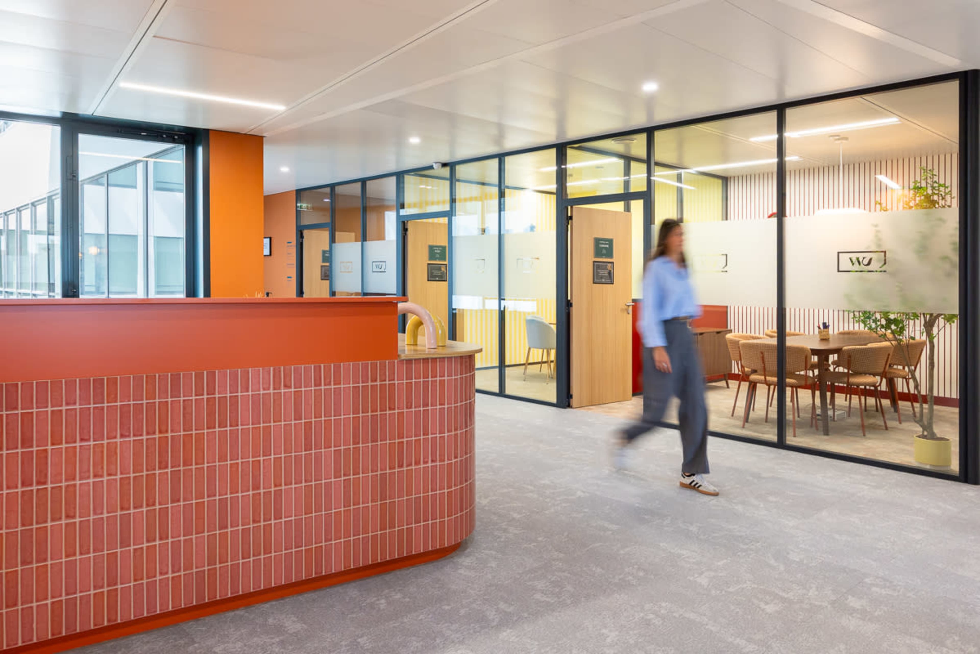 A person walks through a modern office space featuring glass-walled meeting rooms and an orange reception area.