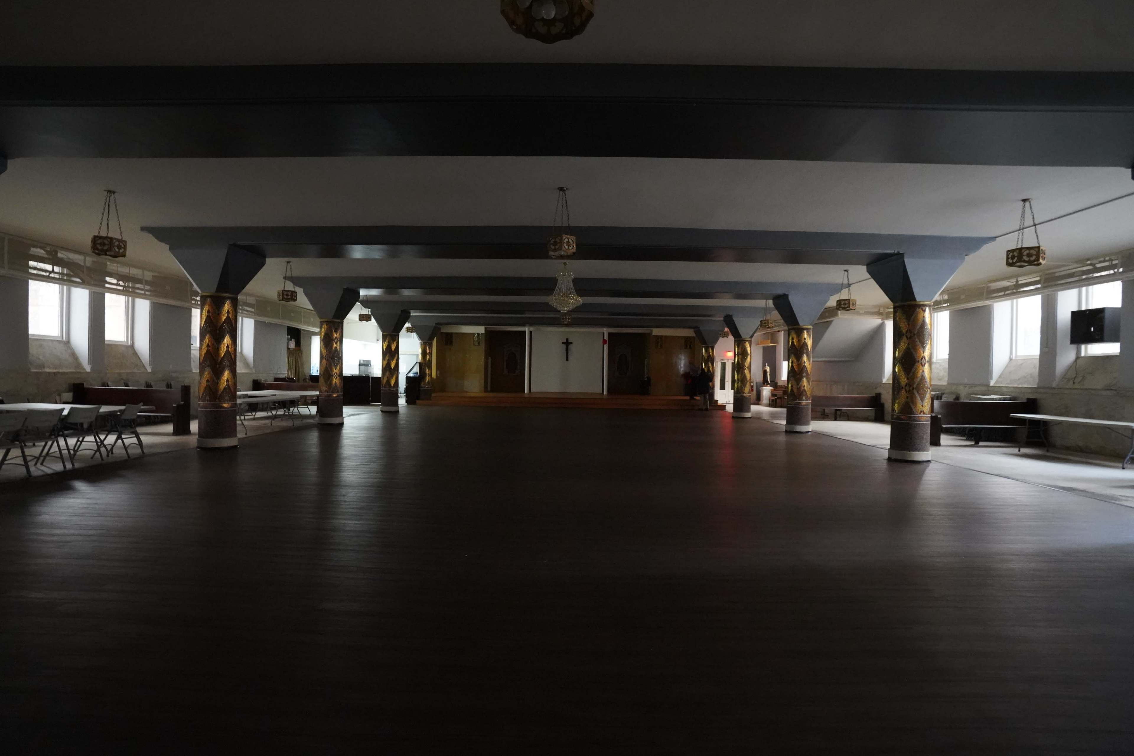 A large, empty hall with polished wooden floors and decorative columns, featuring rows of folding chairs along the sides.
