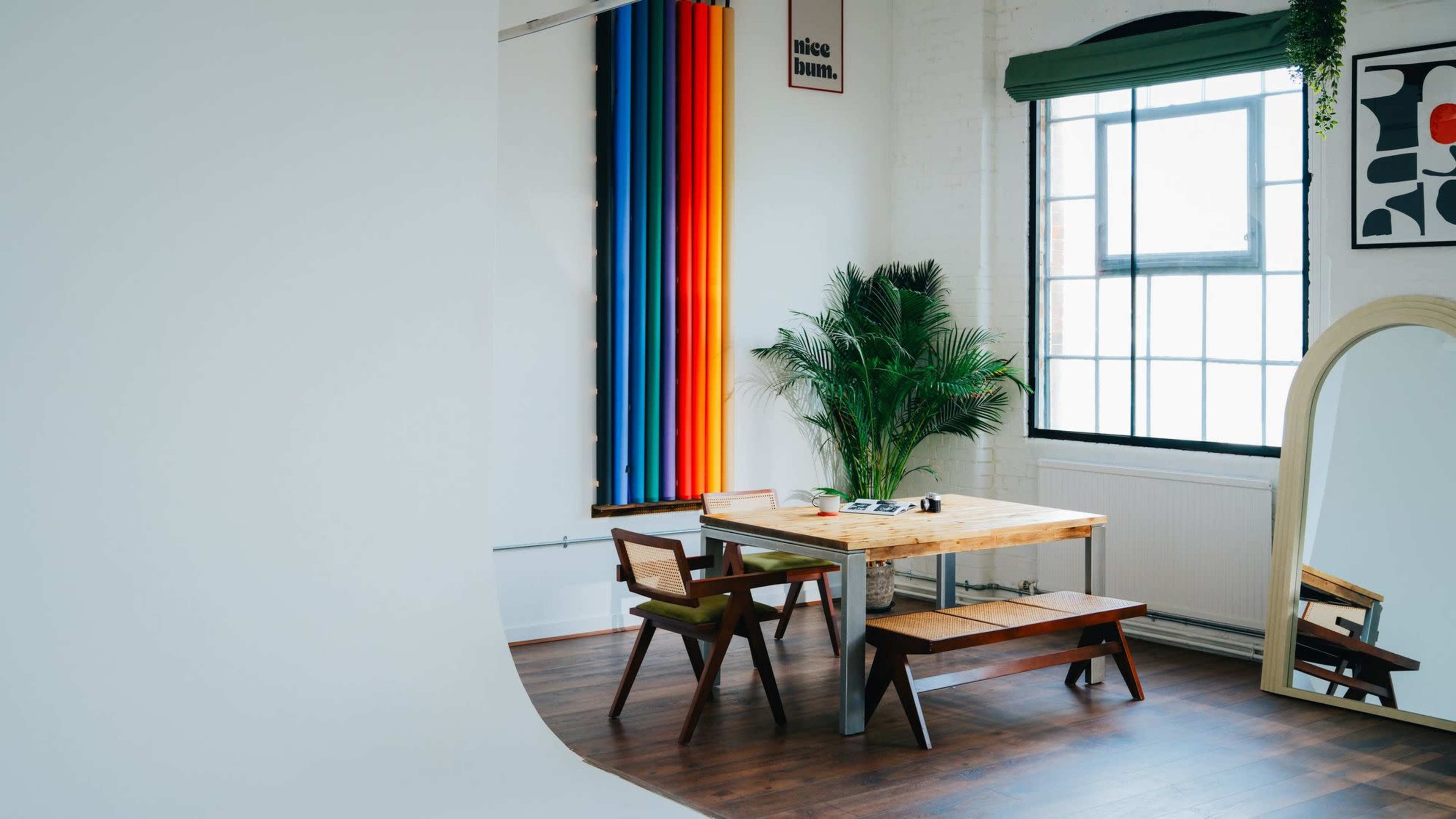 A bright, minimalist studio features a wooden table with benches, colorful backdrop curtains, and a large mirror beside a potted plant.