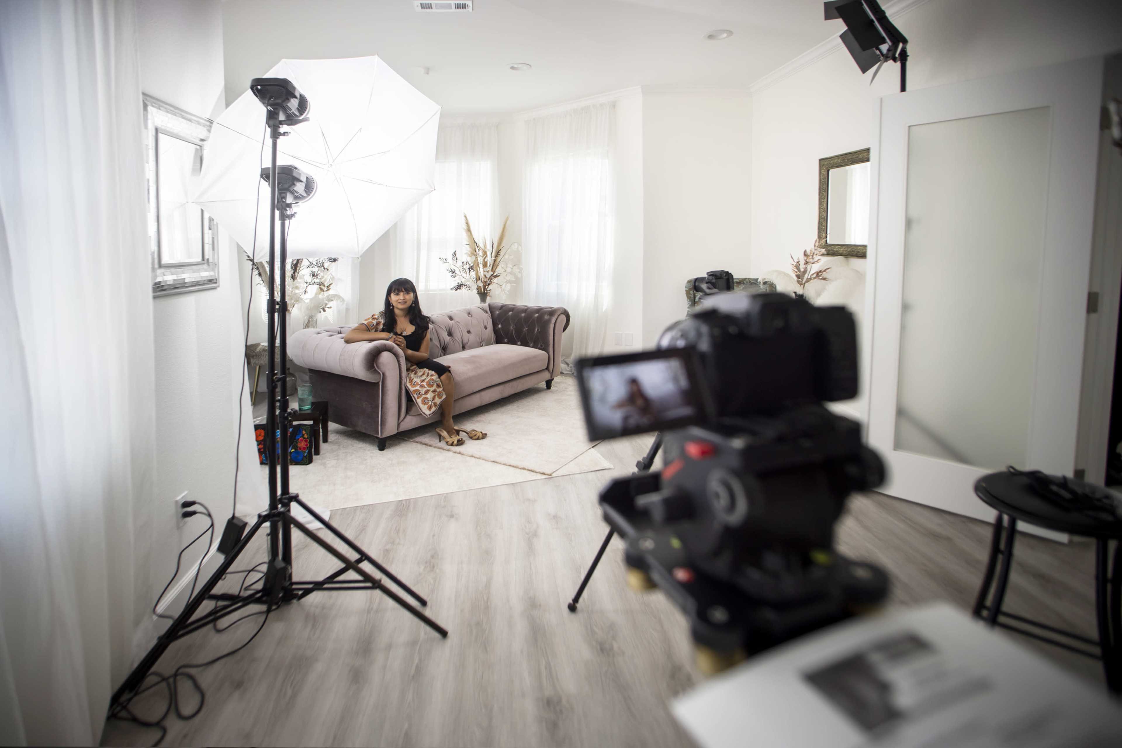 A woman sits on a gray sofa in a well-lit room equipped with photography lighting and a camera on a tripod focused on her.