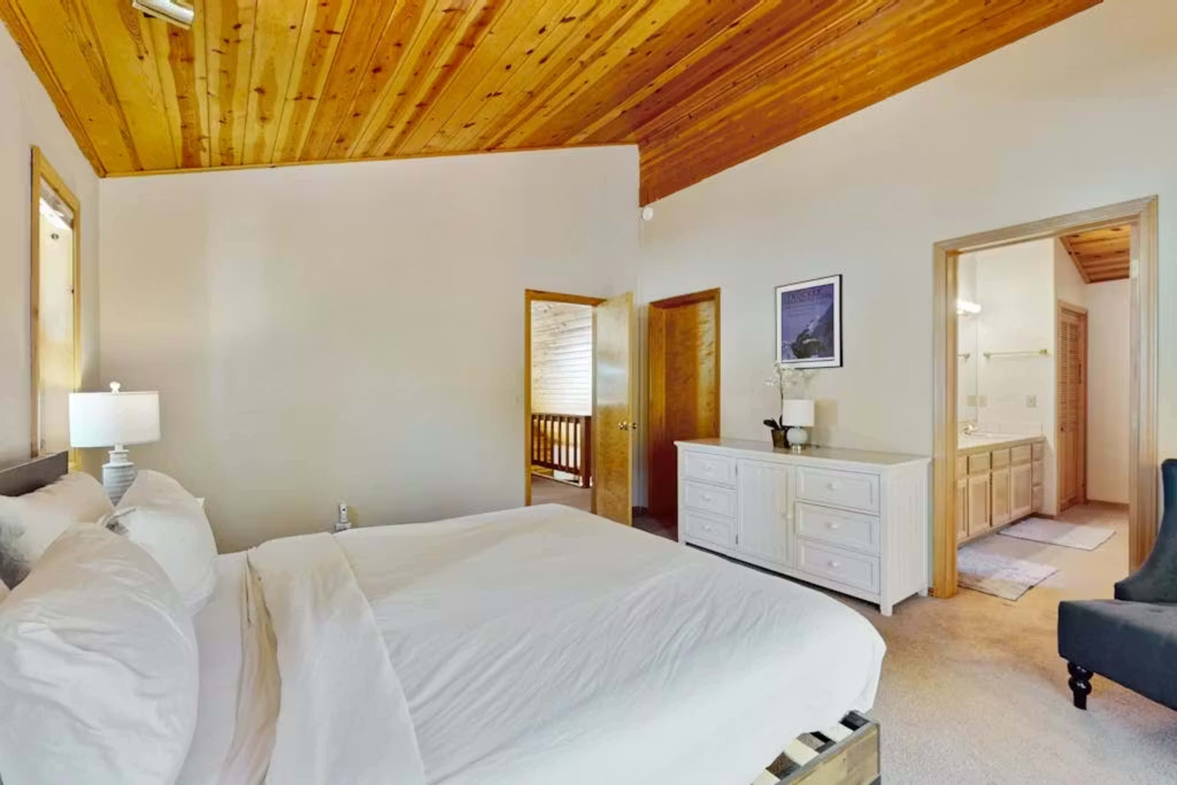 The image shows a cozy bedroom featuring a white bed, wooden ceiling, and adjacent doors leading to other rooms.