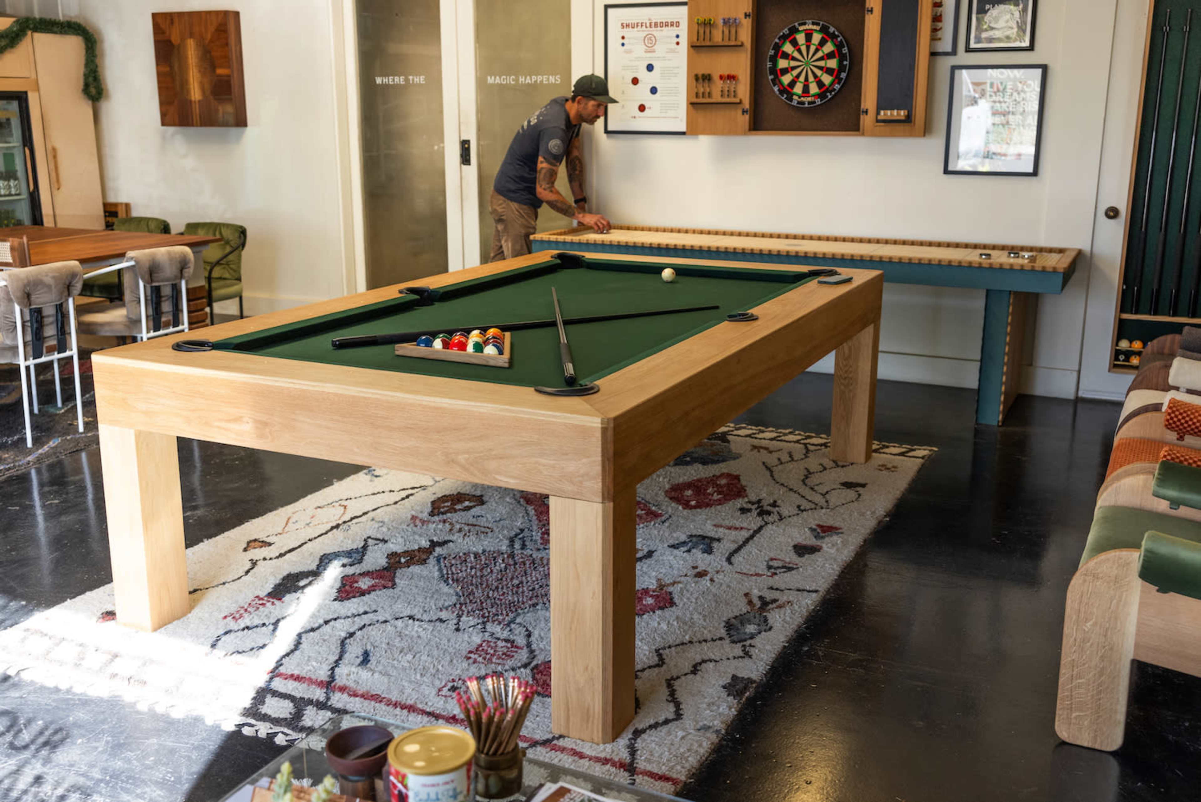 A person is preparing to play pool at a large wooden table in a room with a rug, dartboard, and various decor.