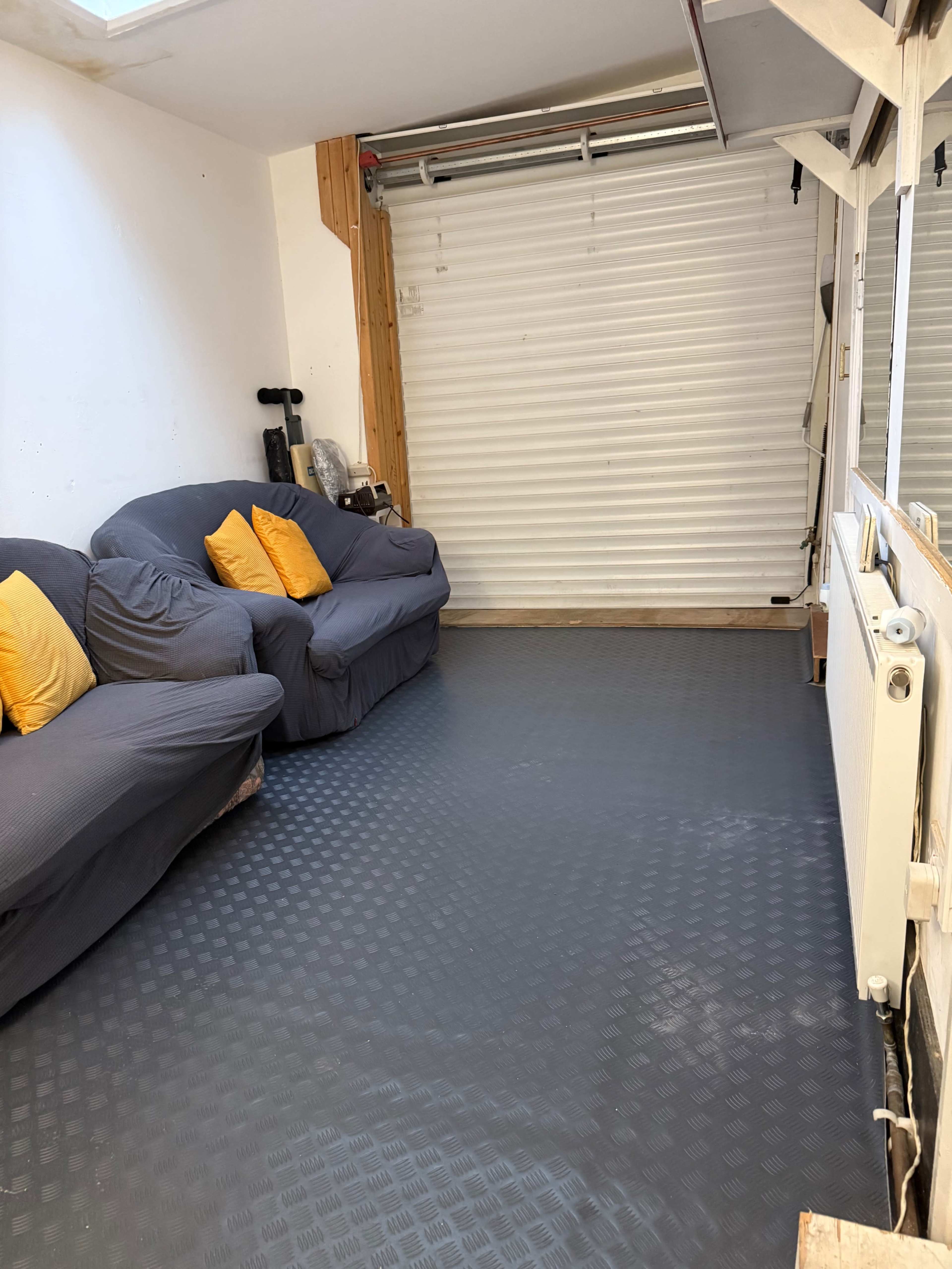 The image shows a room with two gray sofas, each adorned with yellow cushions, facing a closed white roller door and a radiator.