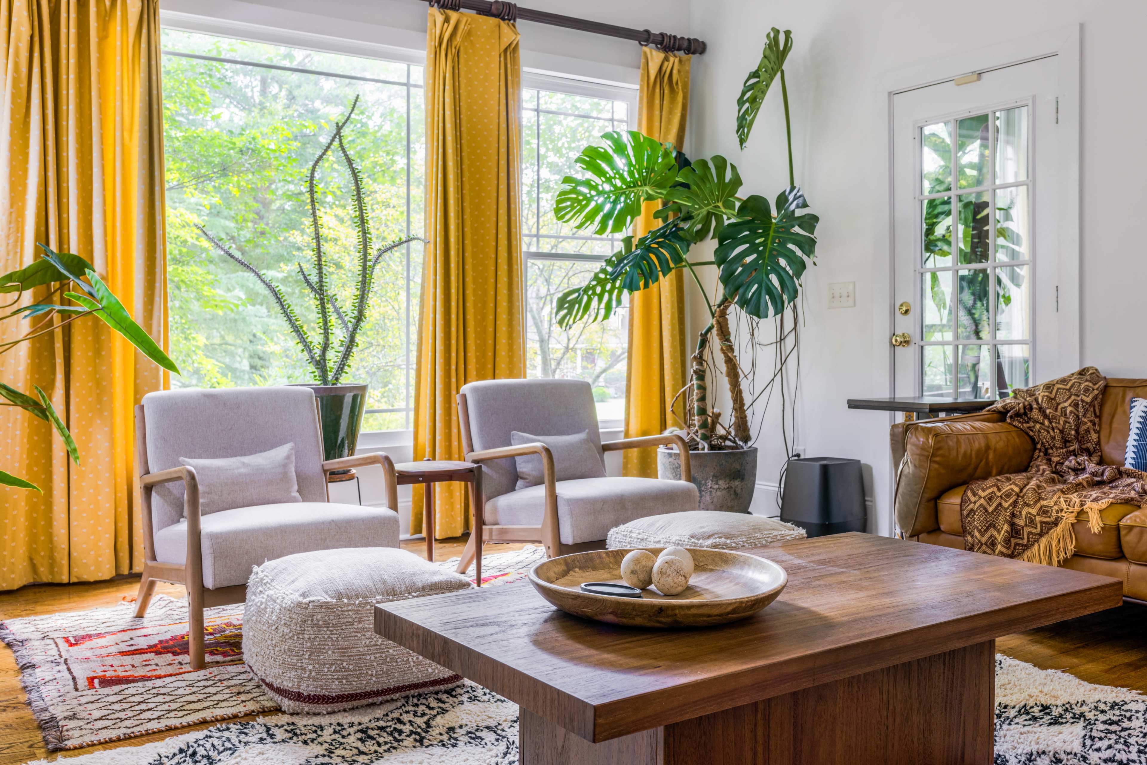 A bright living room with two upholstered chairs, a wooden coffee table, large potted plants, and yellow curtains framing a window.