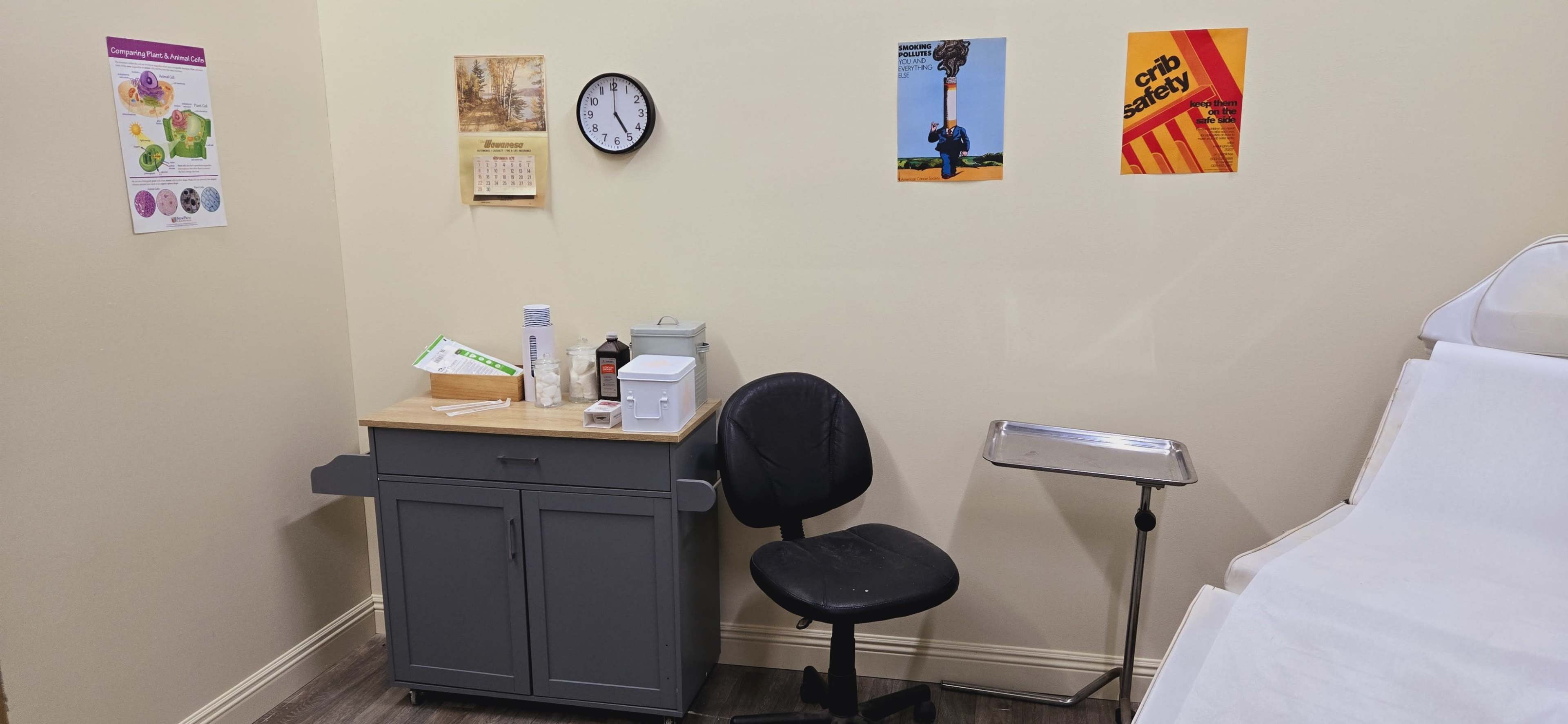 A small room with a clock, a calendar, and safety posters on the walls, along with a desk, a chair, and a medical tray.