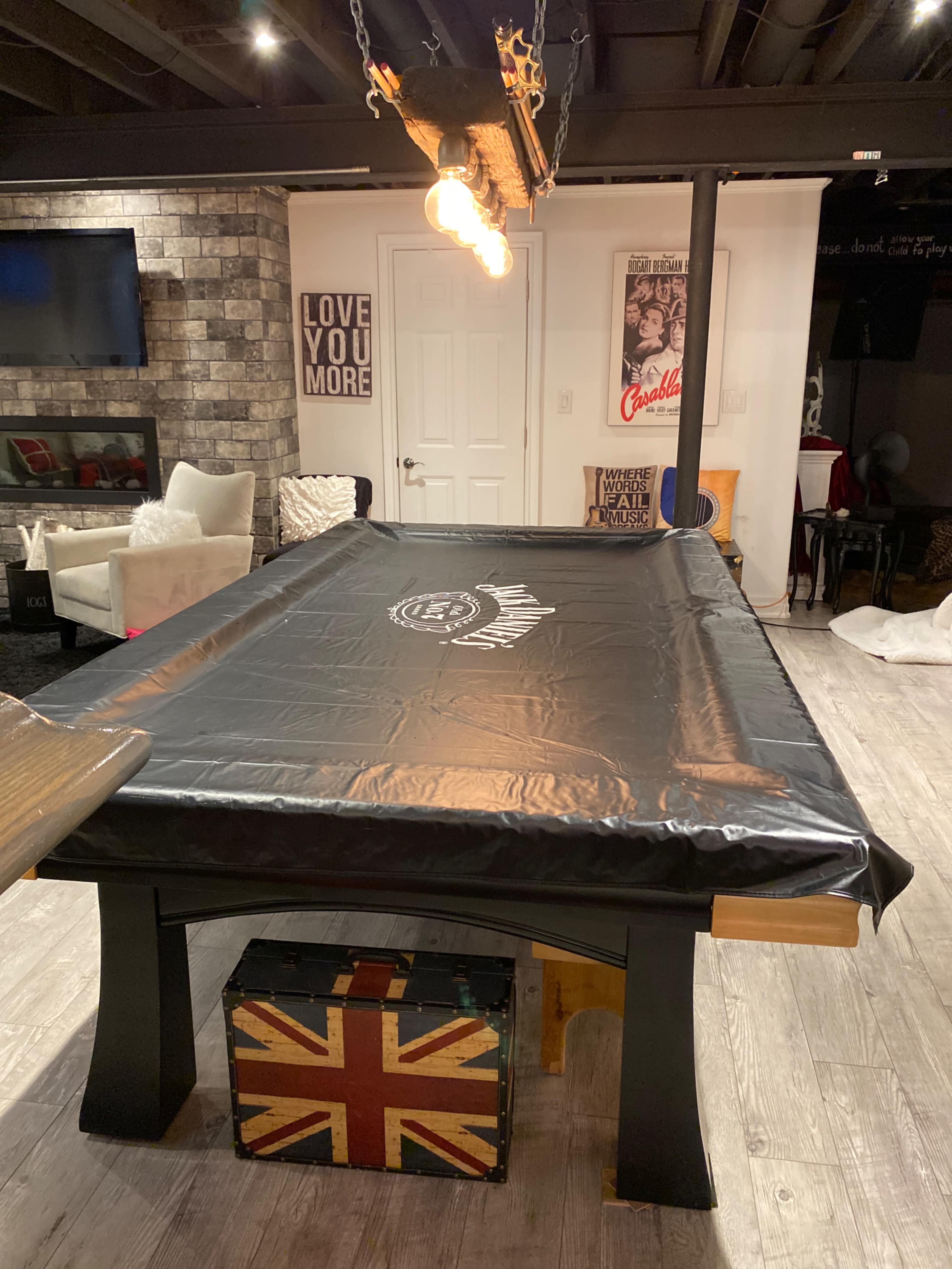 A dimly lit basement room with a large black-covered pool table and a vintage-style trunk with a Union Jack design.