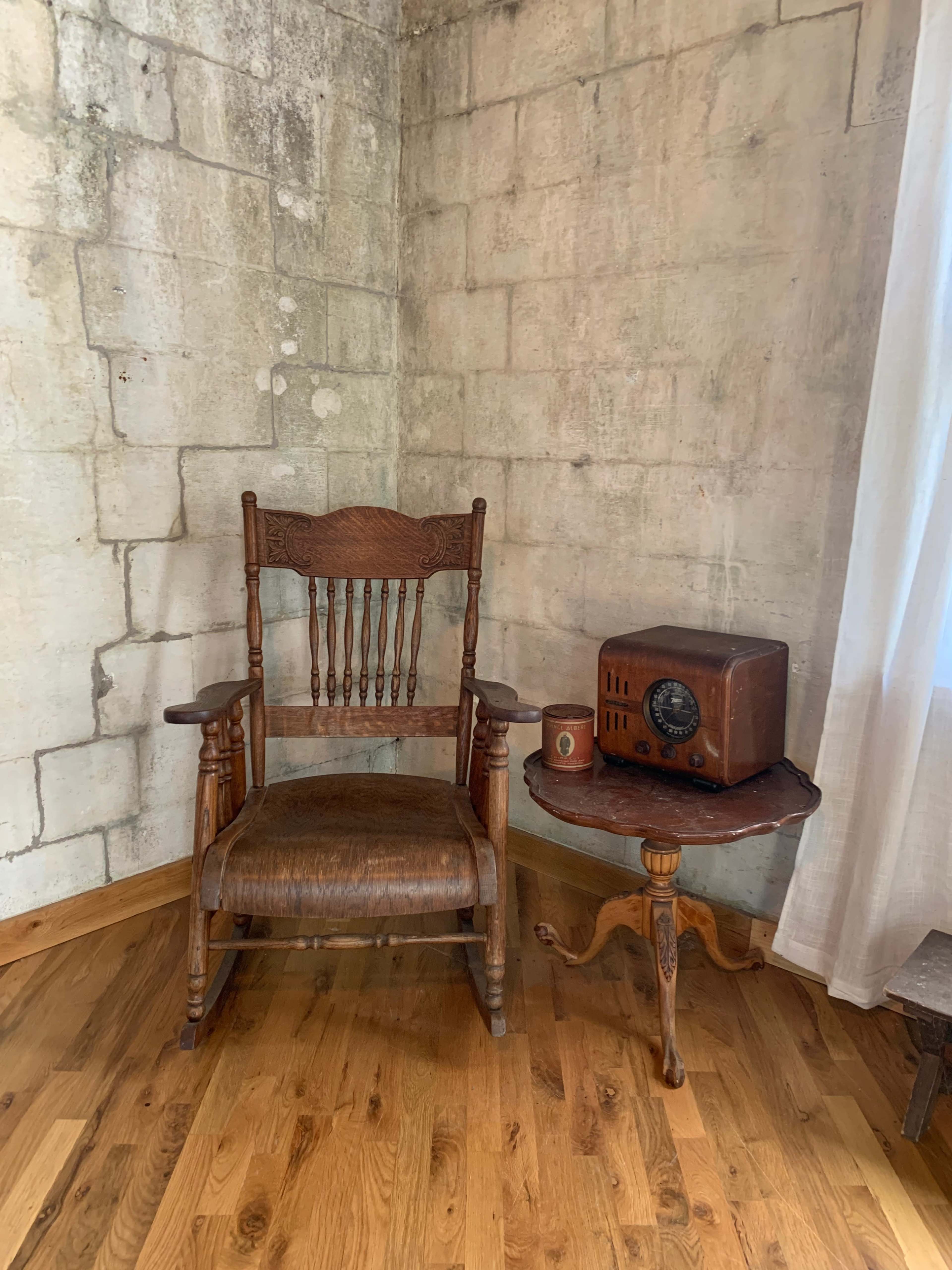 A wooden rocking chair sits beside a small round table with an old-fashioned radio, against a textured stone wall.