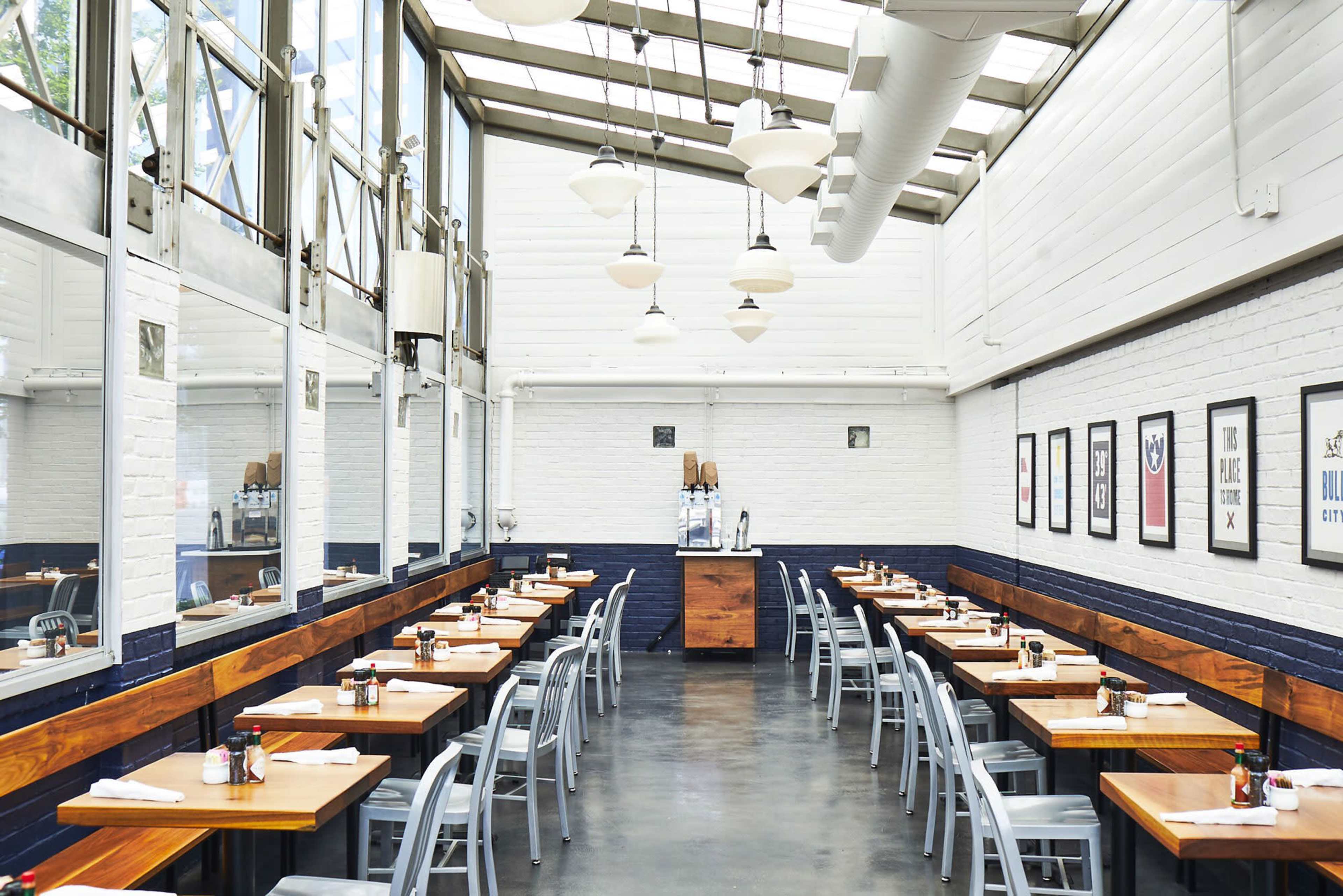 The image shows a bright, modern restaurant featuring rows of wooden tables and gray chairs, with large windows allowing natural light and decorative artwork on the walls.