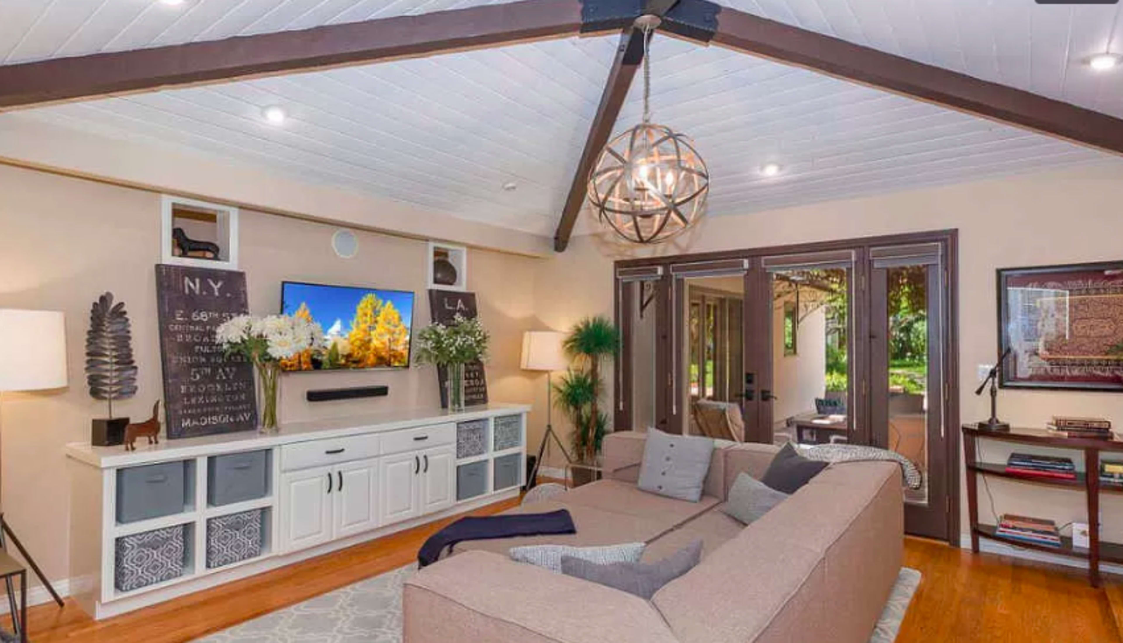 The living room features a sectional sofa, a large television mounted on the wall, and glass doors leading to an outdoor area, all under a vaulted ceiling with exposed beams.