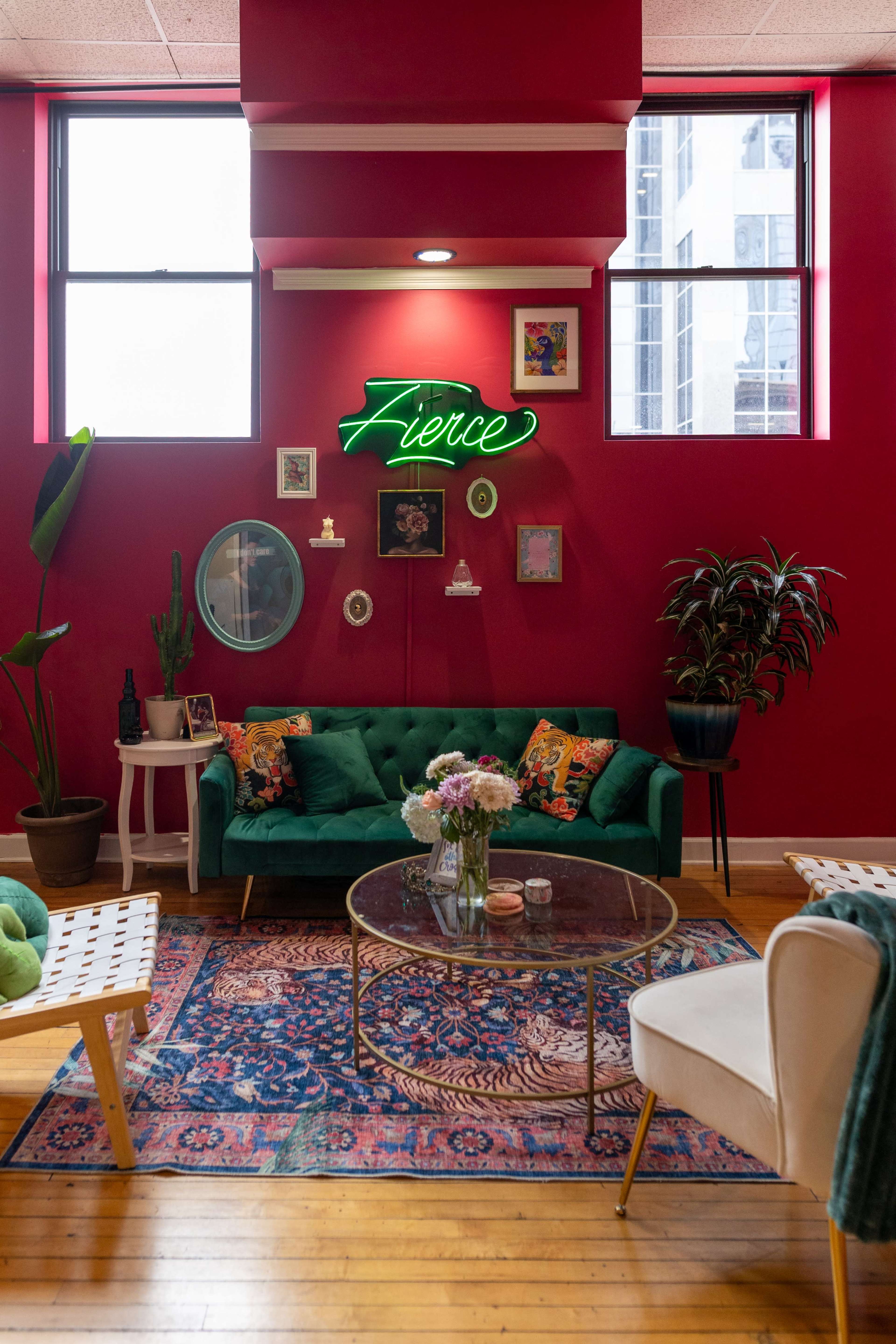 The image shows a cozy, brightly colored lounge area with a green velvet sofa, a glass coffee table, and decorative plants against a vibrant red wall adorned with various framed artworks and a neon sign that says "Fierce."