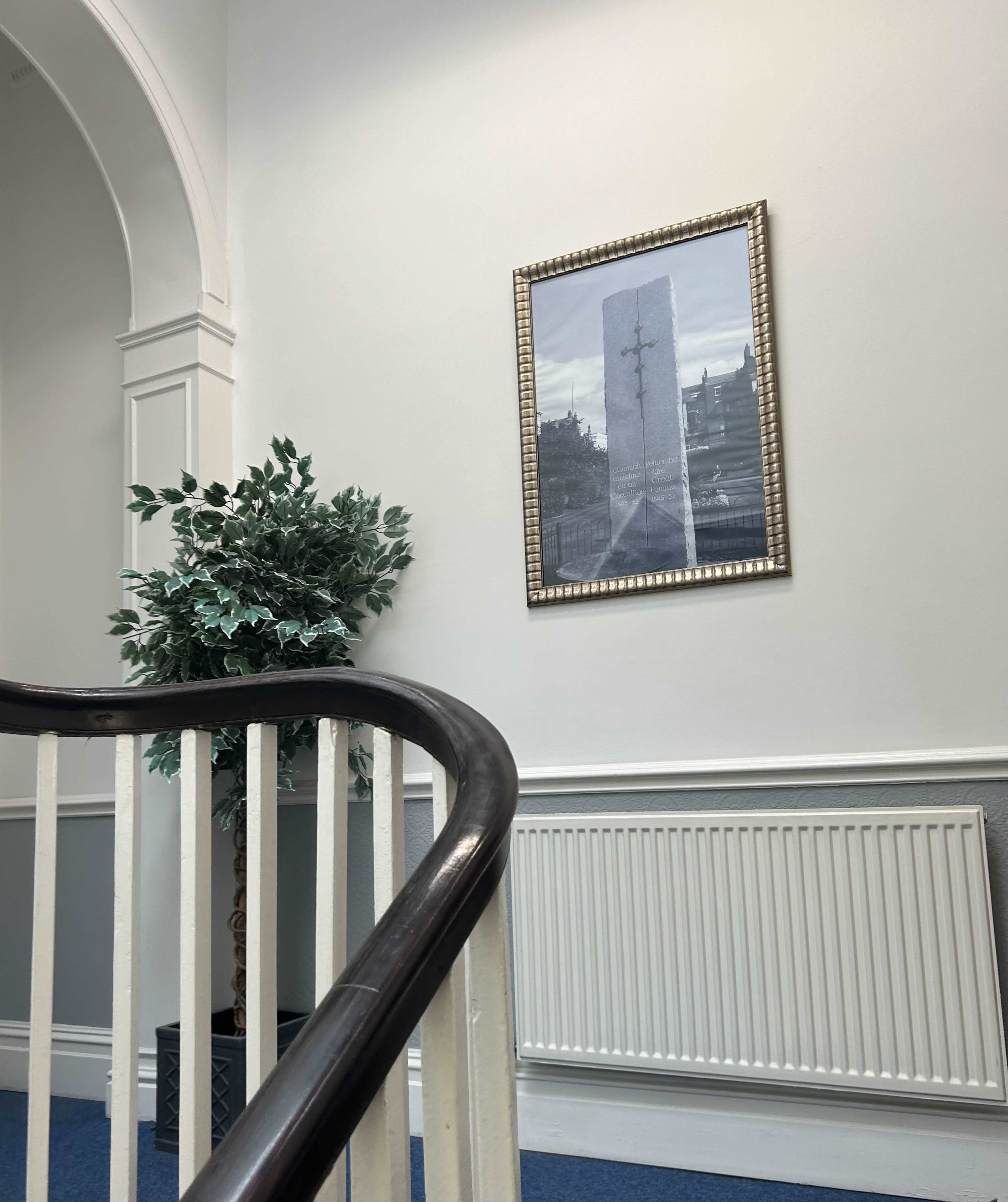 A staircase with a wooden railing leads to a wall displaying a framed black-and-white photograph of a monument, alongside a tall potted plant.