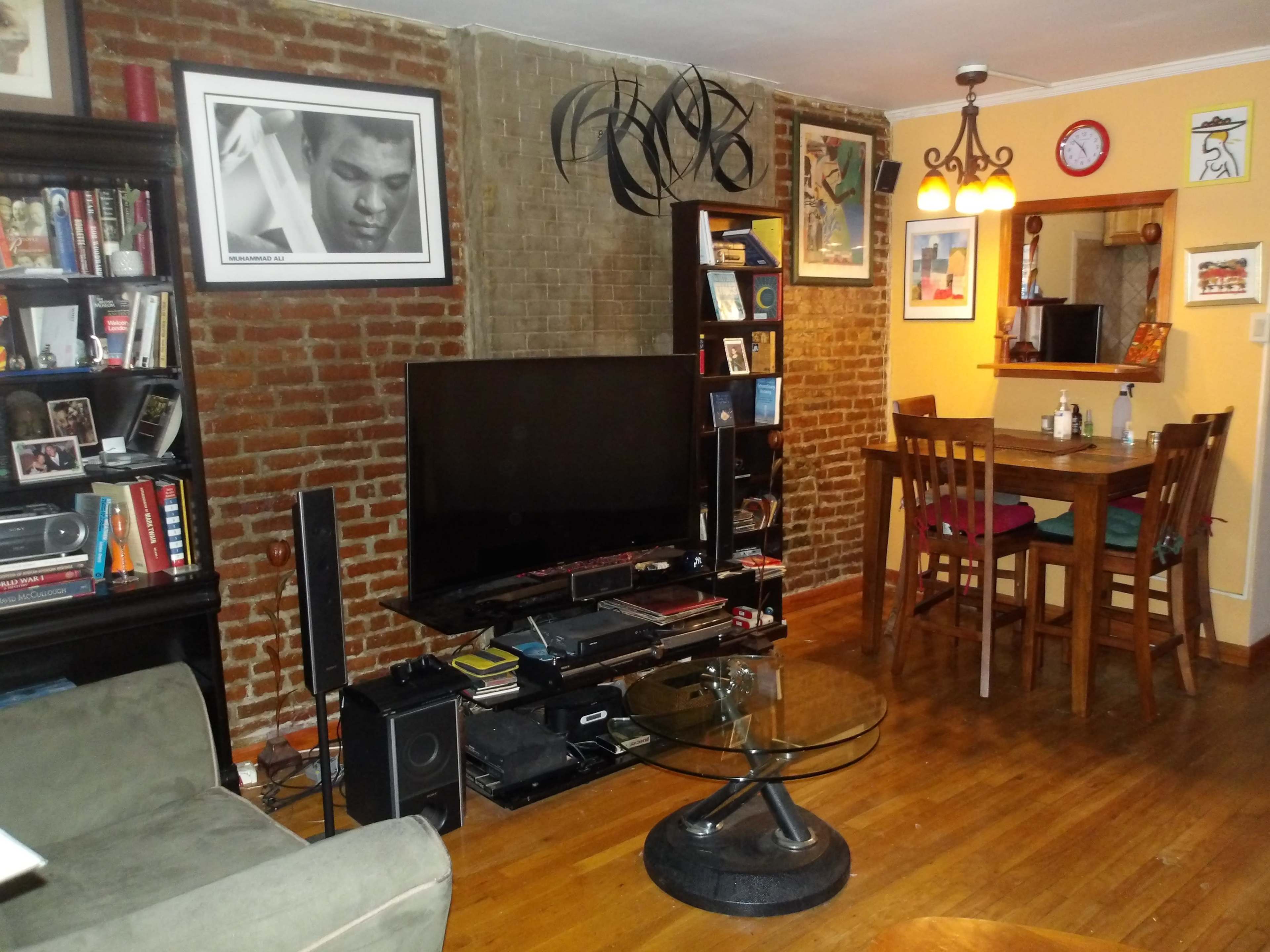 A cozy living room with exposed brick walls, a large TV on a stand, a bookshelf filled with books, a circular glass coffee table, and a dining area with a wooden table and chairs.