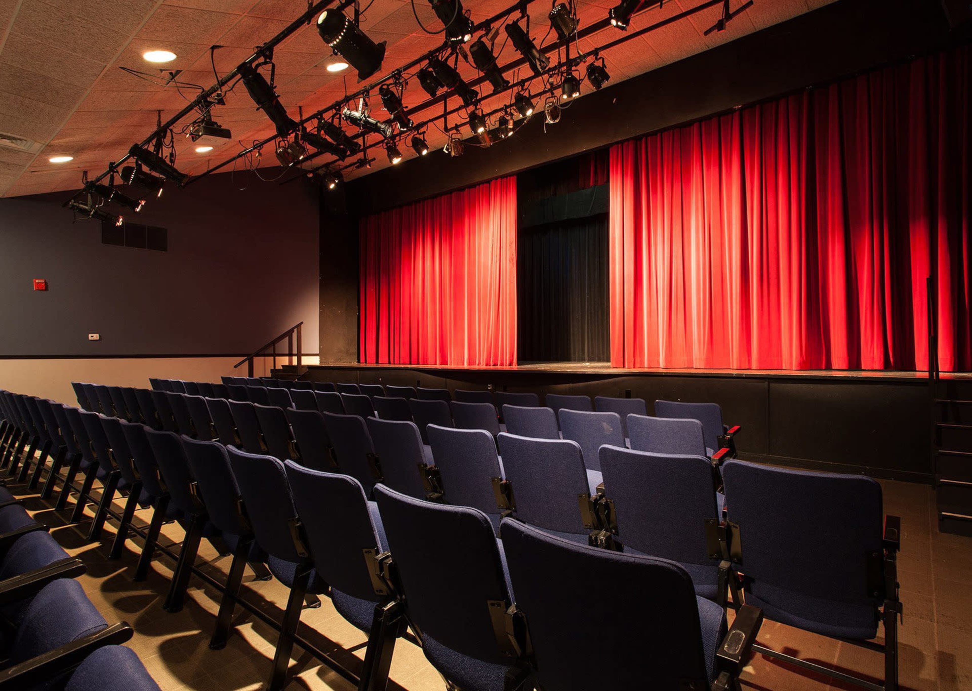 A theater with blue chairs arranged in rows facing a stage adorned with red curtains and overhead lighting.