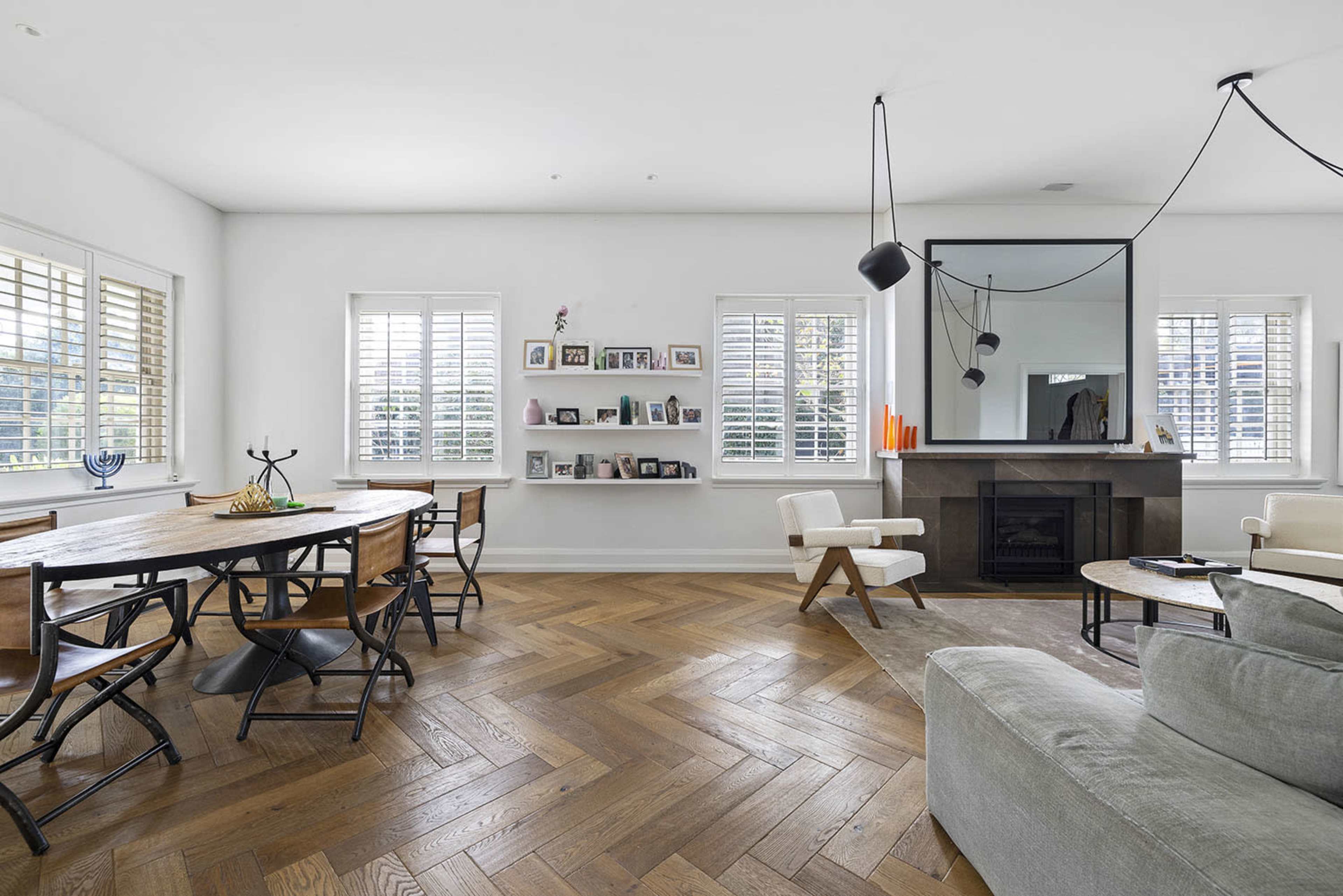 A spacious, modern living area with a dining table, a fireplace, and large windows with shutters.