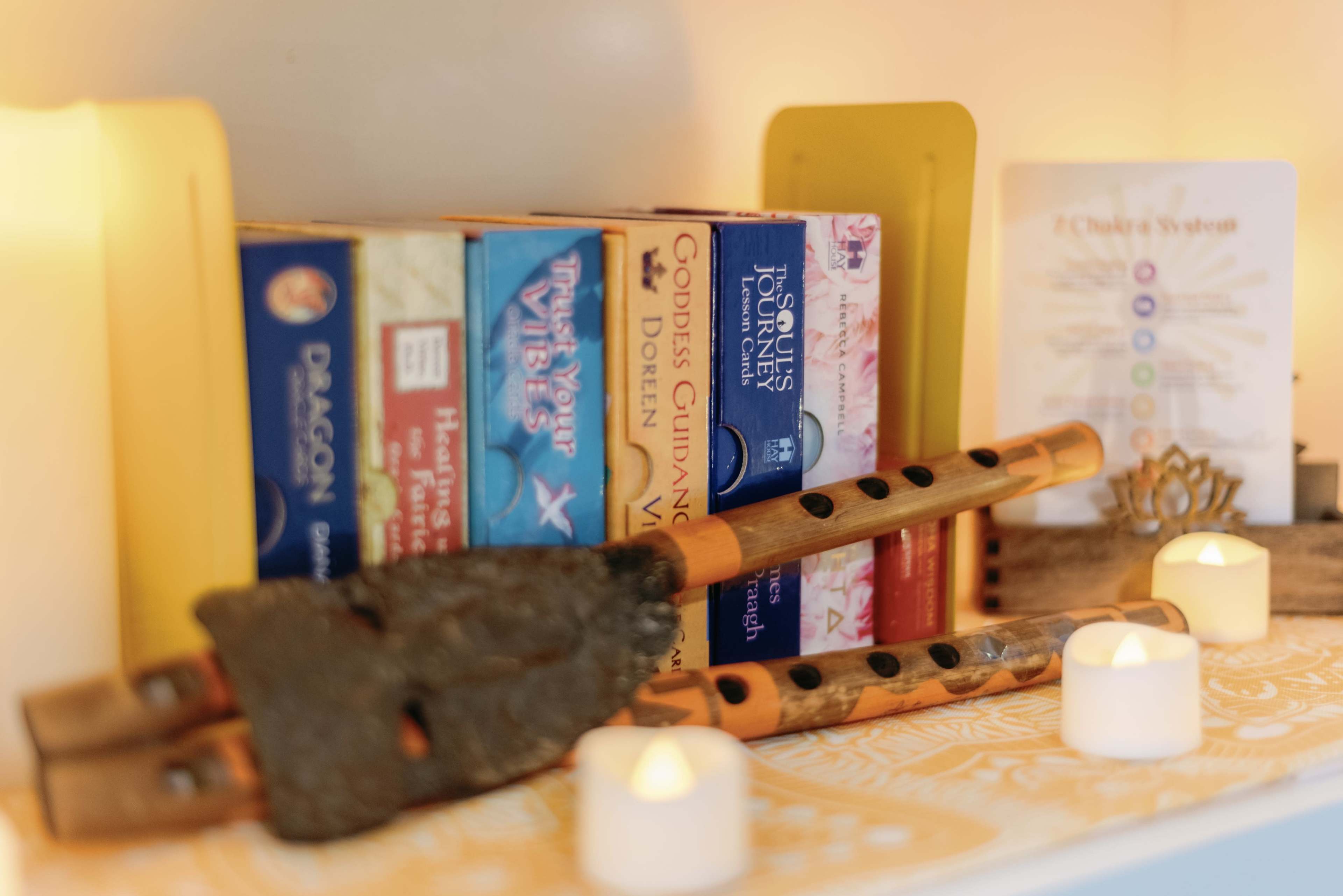 The image shows a shelf with several colorful boxes of cards, two wooden flutes, a small decorative item, and lit candles arranged on a patterned cloth.