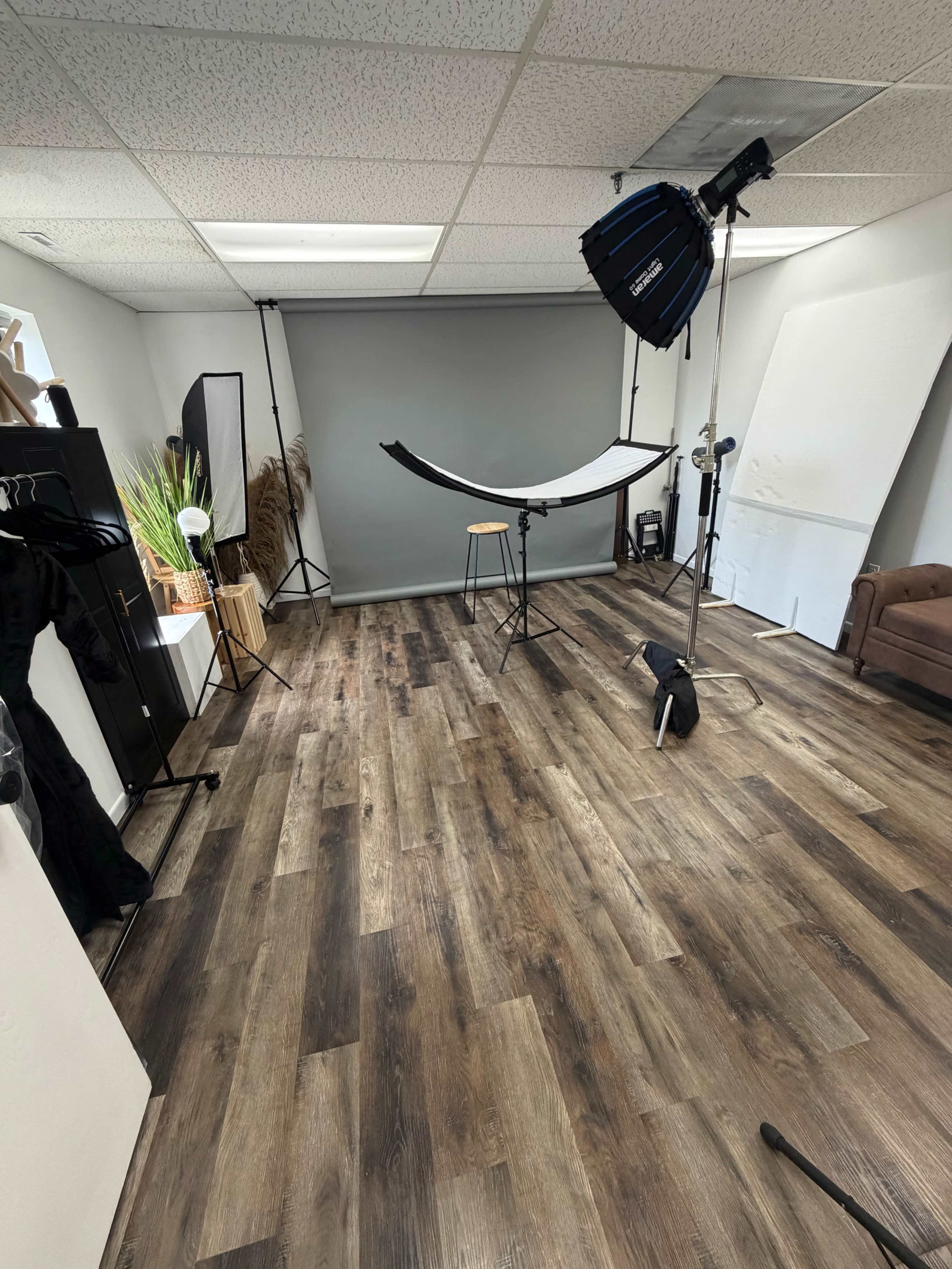 The image shows a photography studio with wooden floors, a backdrop setup, lighting equipment, and a brown sofa.