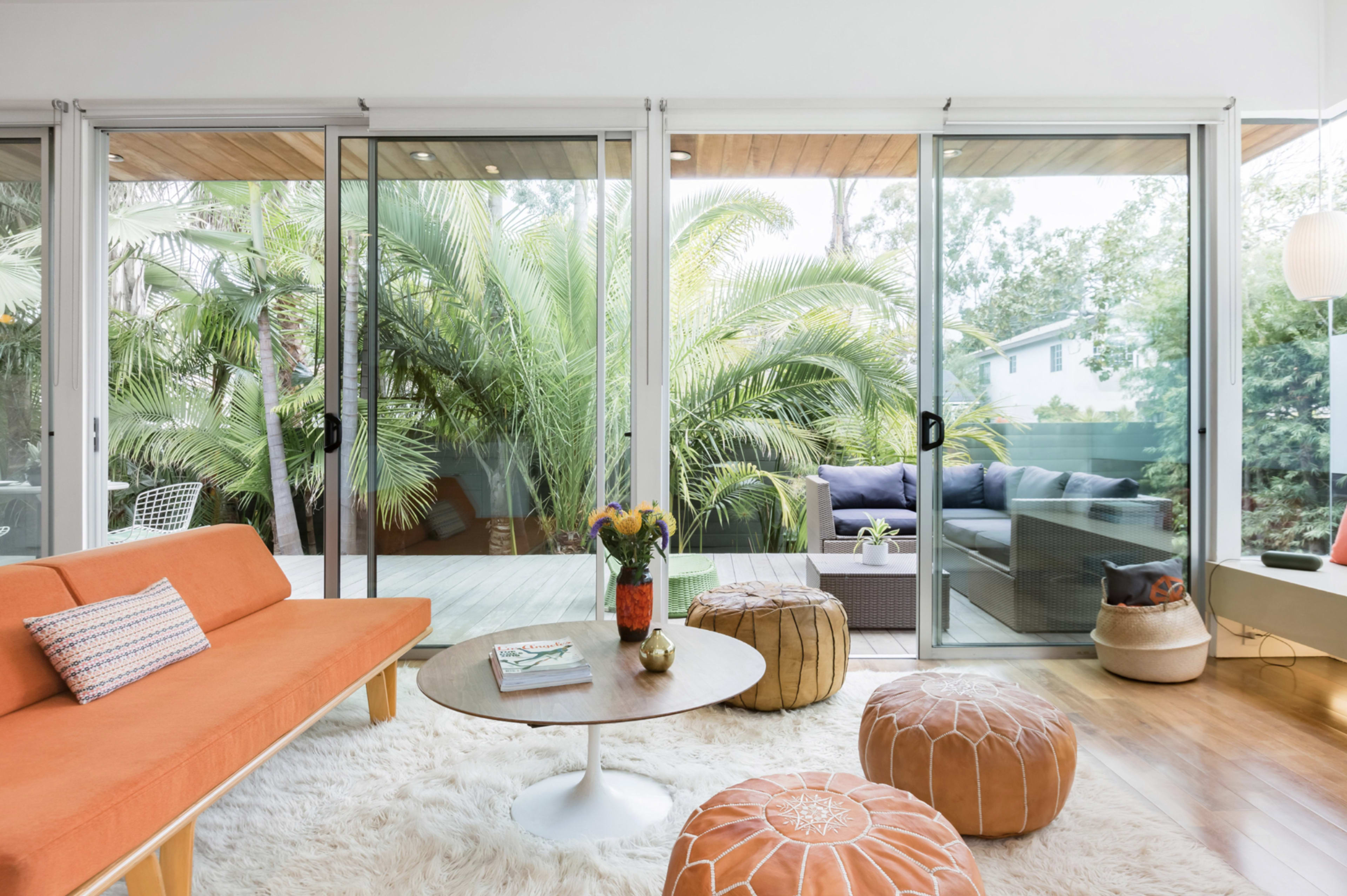 The image features a bright, modern living room with an orange sofa, a round coffee table, and large glass doors that open to a lush outdoor area filled with palm trees.
