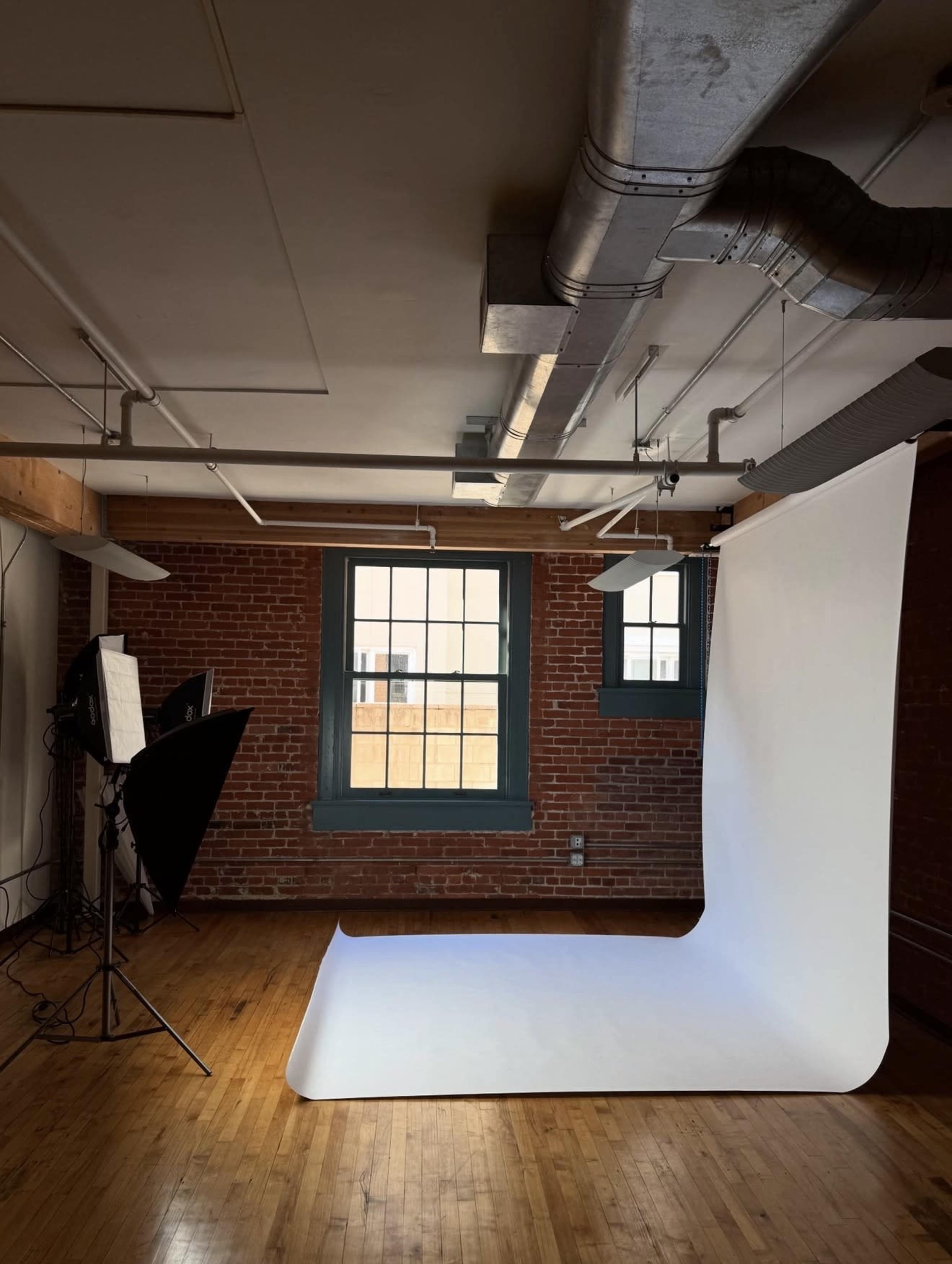 The image shows a photography studio with a white backdrop, wooden floors, and exposed brick walls, illuminated by studio lights.