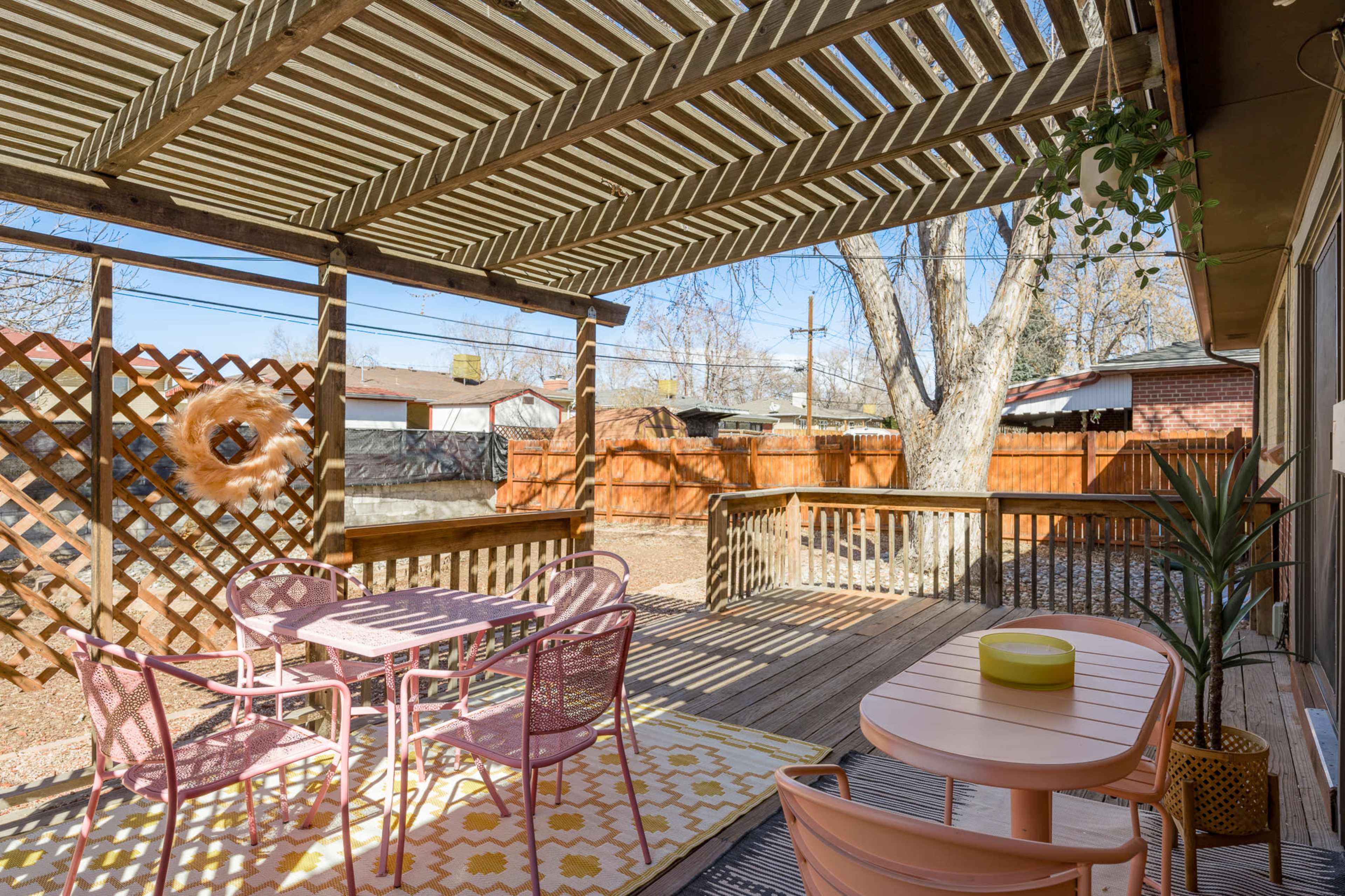 The image shows a wooden patio area featuring a table and chairs under a slatted roof, with a patterned outdoor rug and a potted plant.