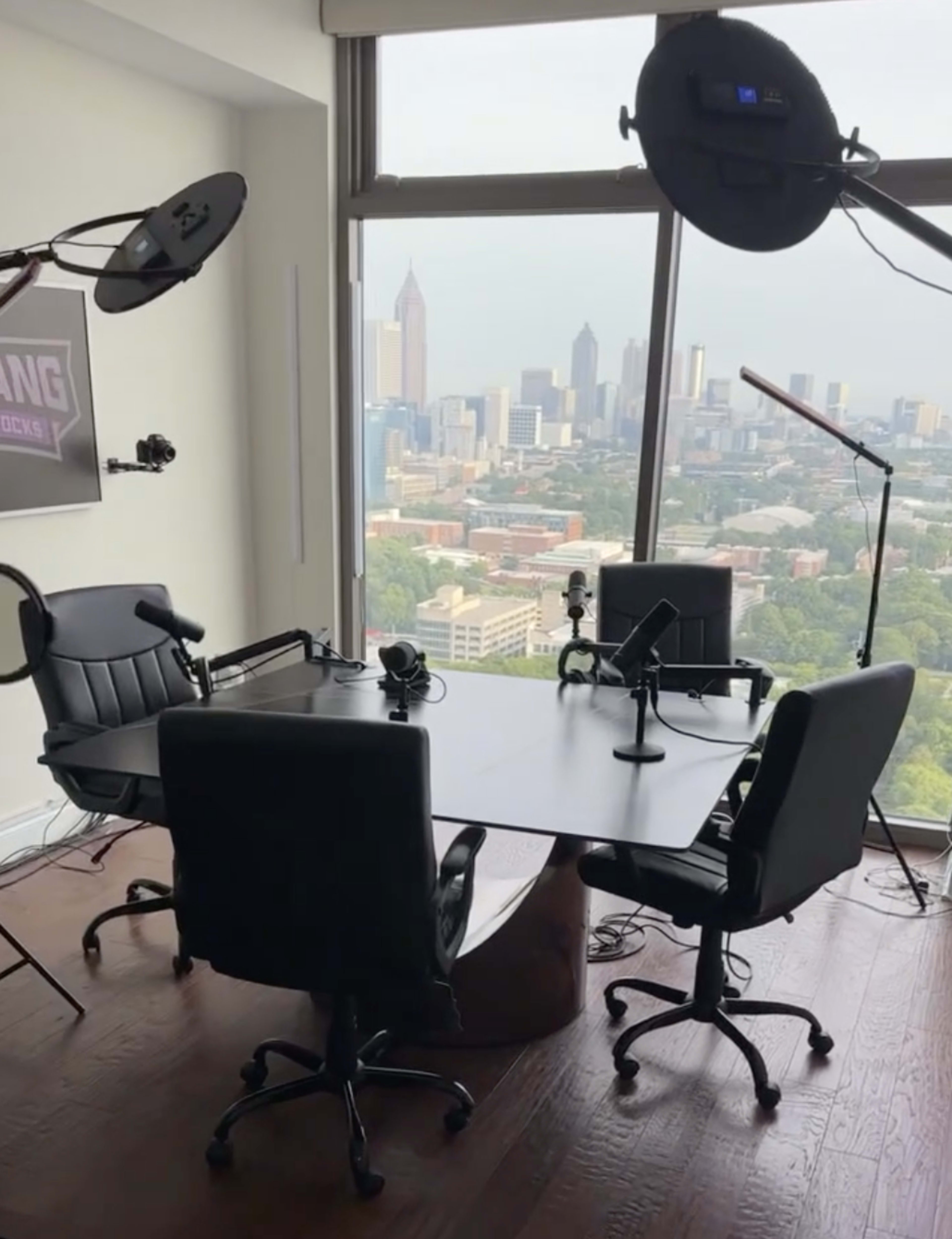 A modern podcast studio with a round table, four black chairs, microphones, and large windows offering a view of a city skyline.