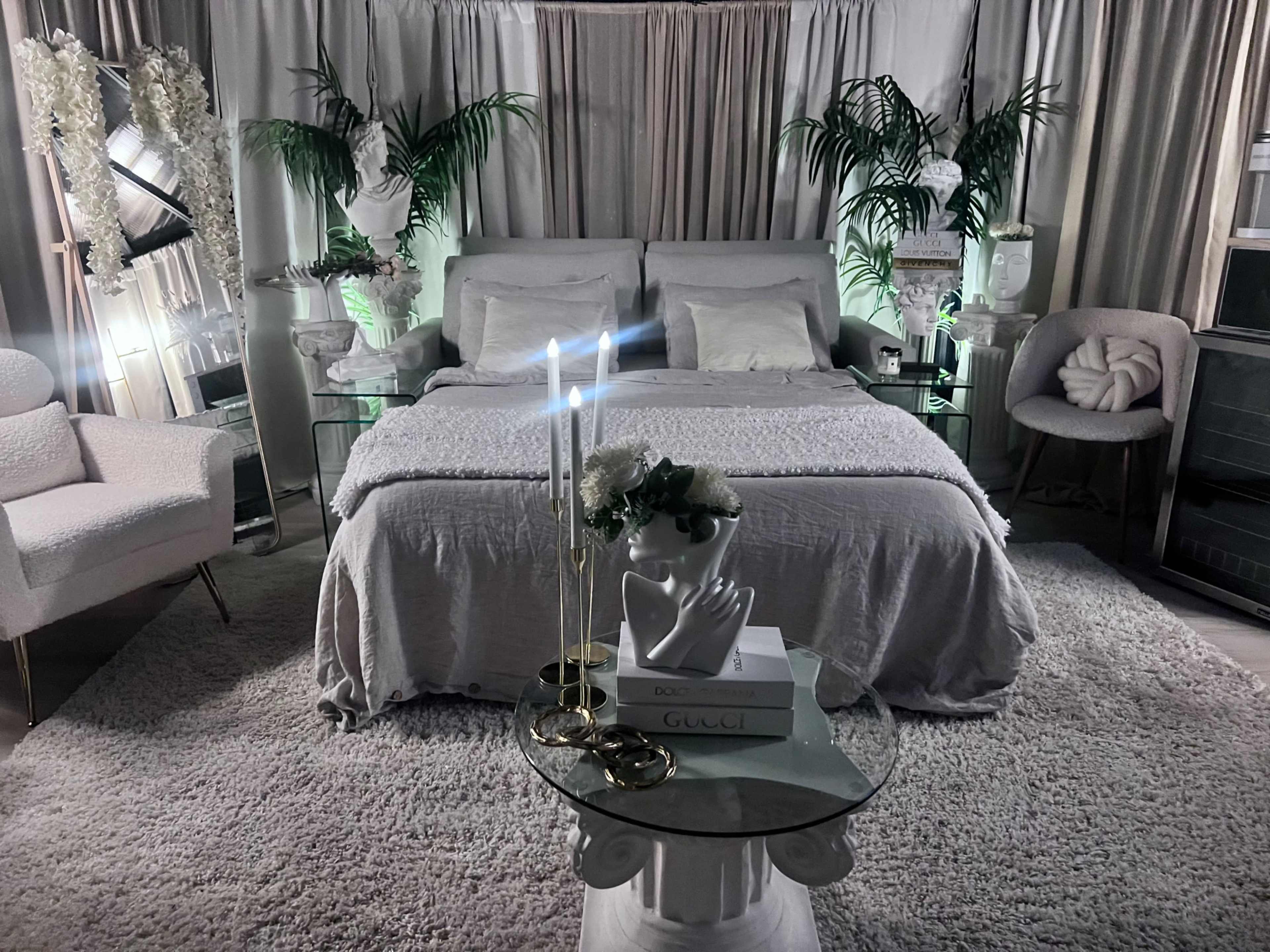 A modern bedroom featuring a large bed with light bedding, decorative plants, and minimalist furniture including a small coffee table and lounge chair.