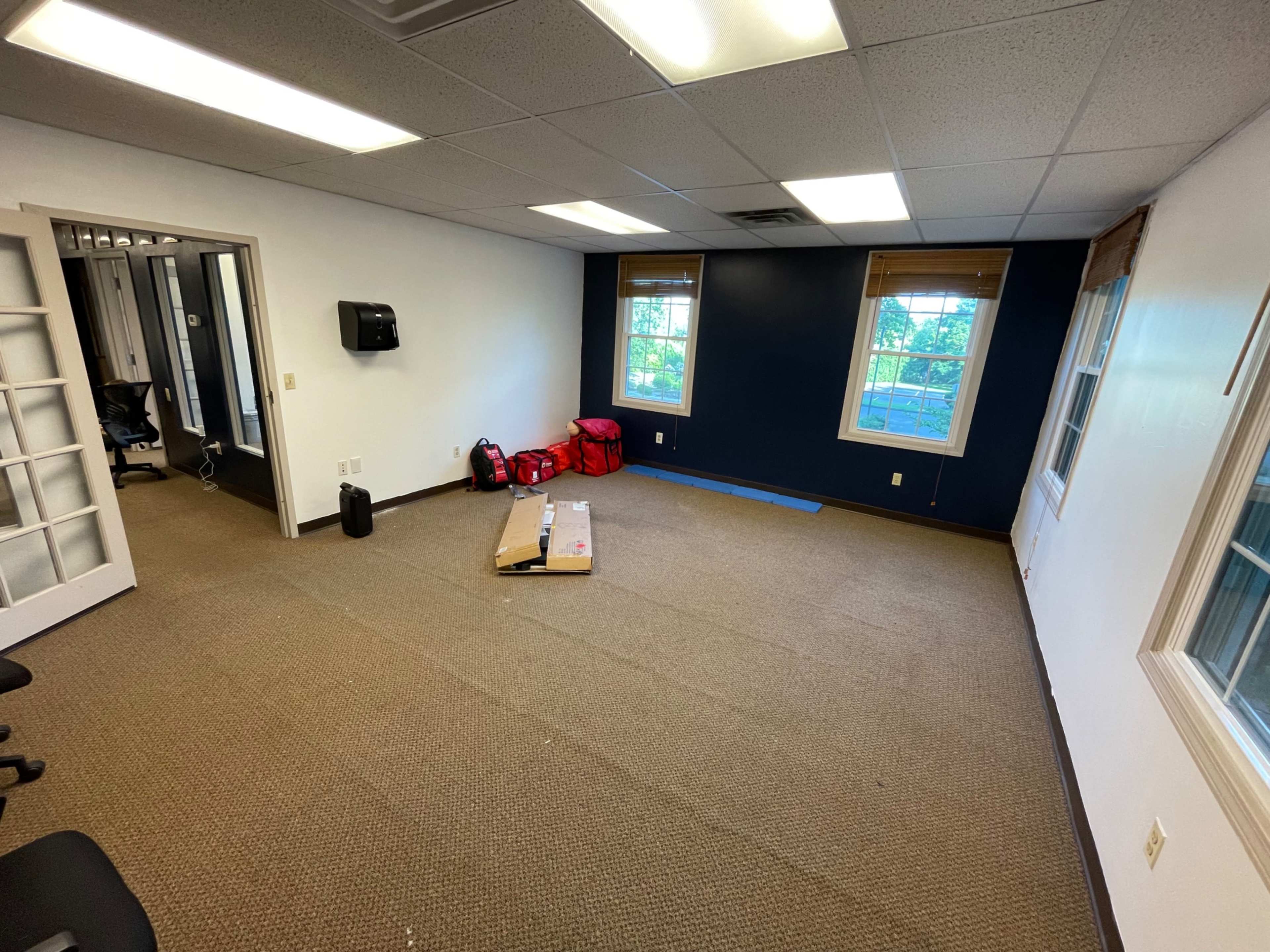 The image shows an empty office space with brown carpeting, two windows, and some scattered items, including red bags and a cardboard box.