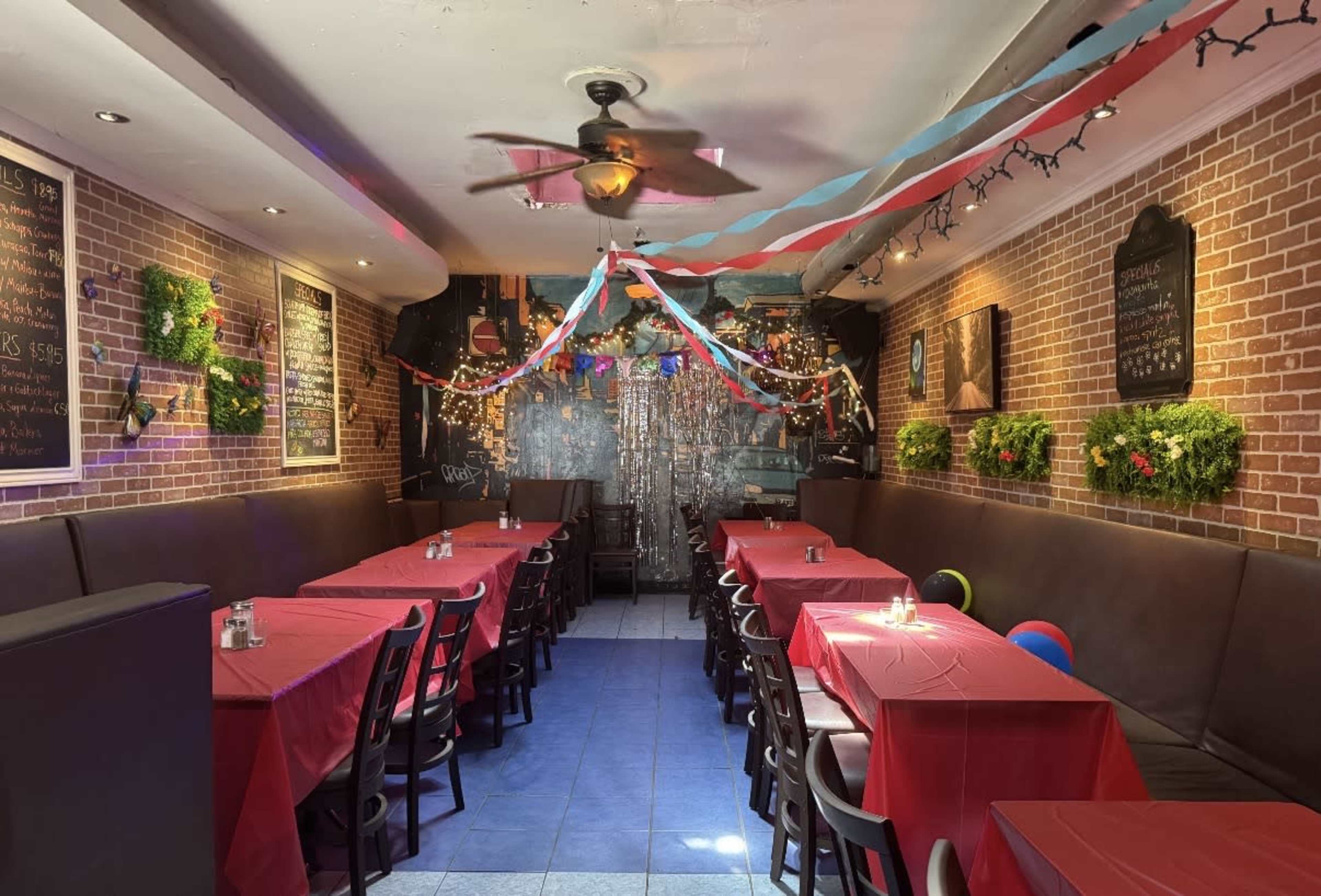 A narrow restaurant space features long tables covered with red tablecloths, surrounded by brick walls adorned with green plants and festive decorations.