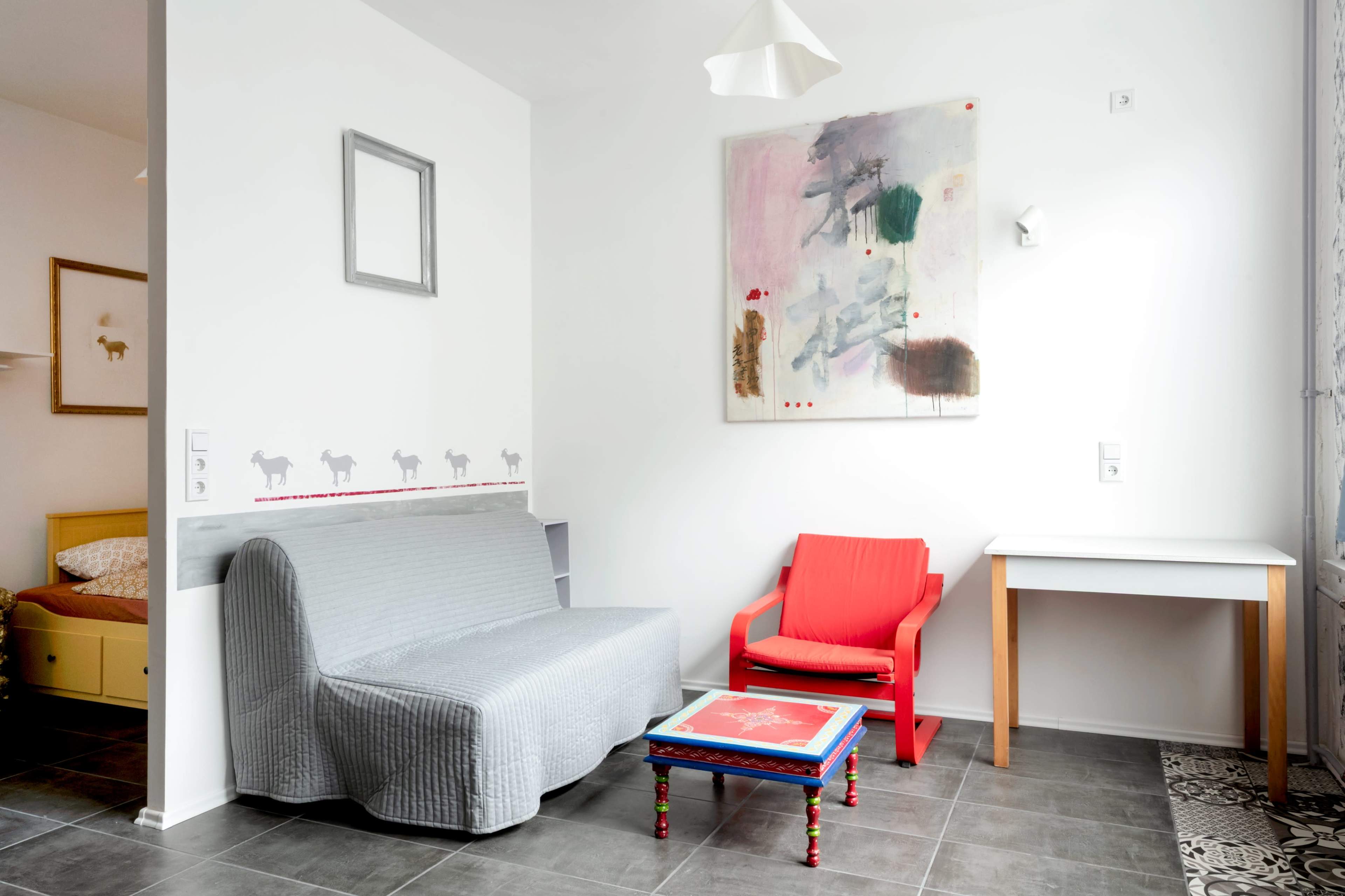 The image shows a modern living room featuring a gray sofa, a red chair, and a colorful coffee table under a light fixture.