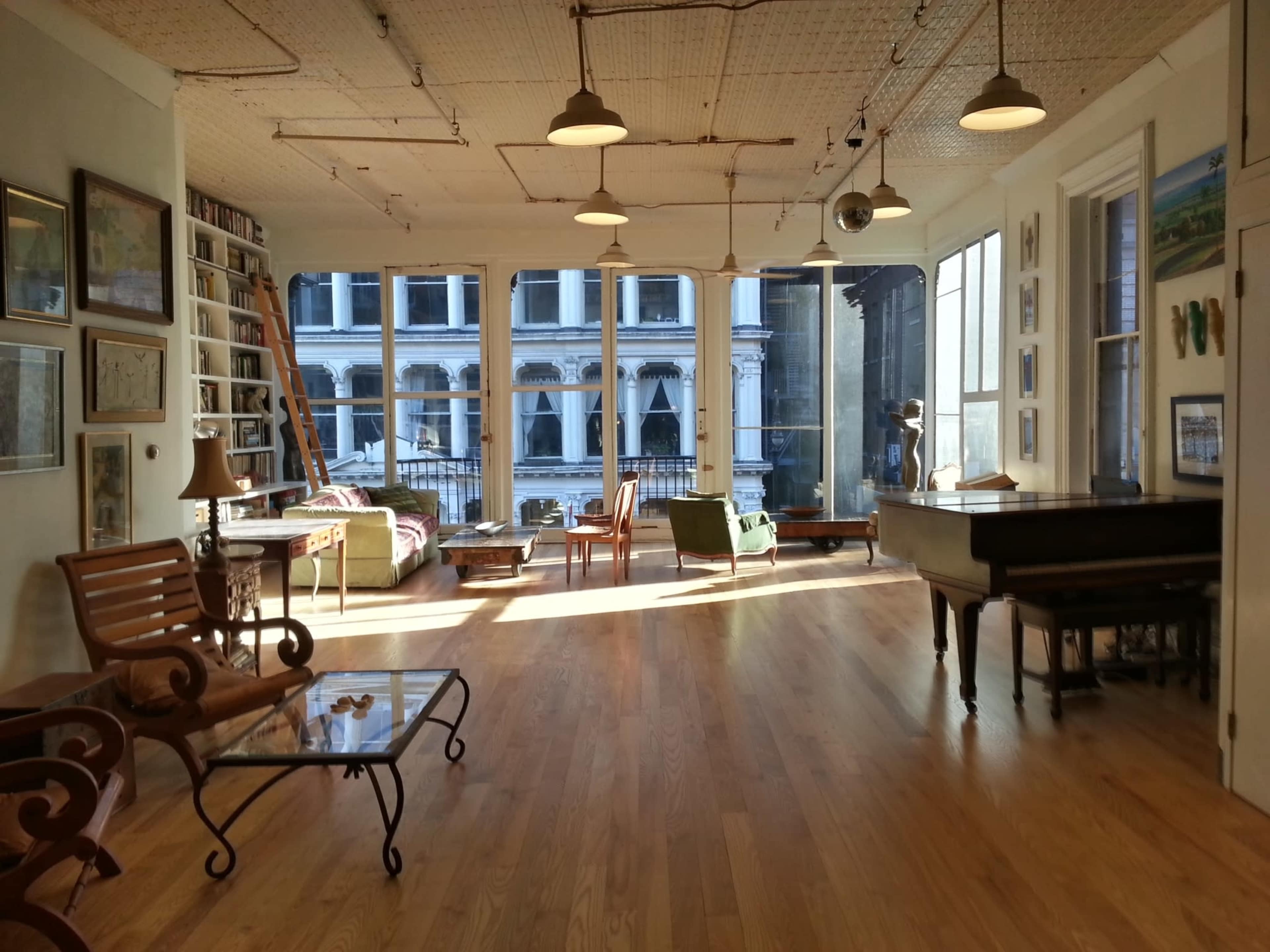A bright, spacious living area with large windows, wooden floors, and various pieces of furniture, including a grand piano and a comfortable seating arrangement.