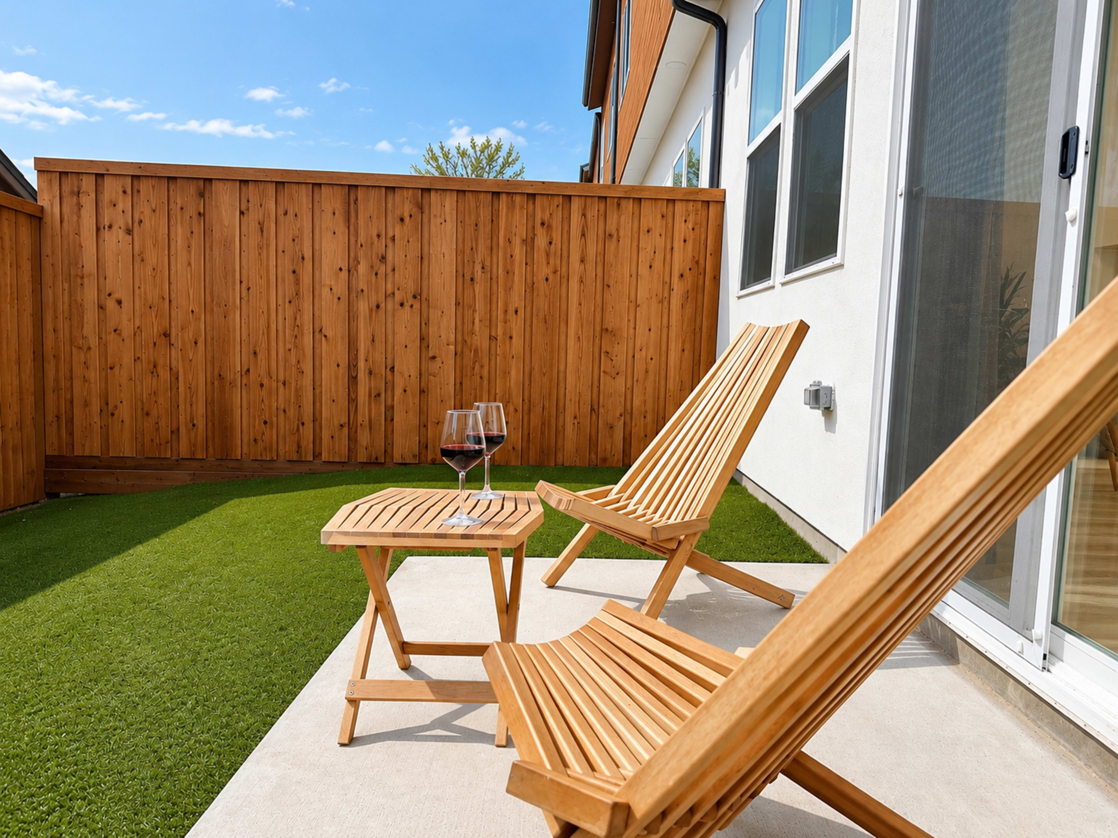A small outdoor patio features two wooden lounge chairs and a side table with two glasses of red wine, surrounded by a wooden fence and artificial grass.