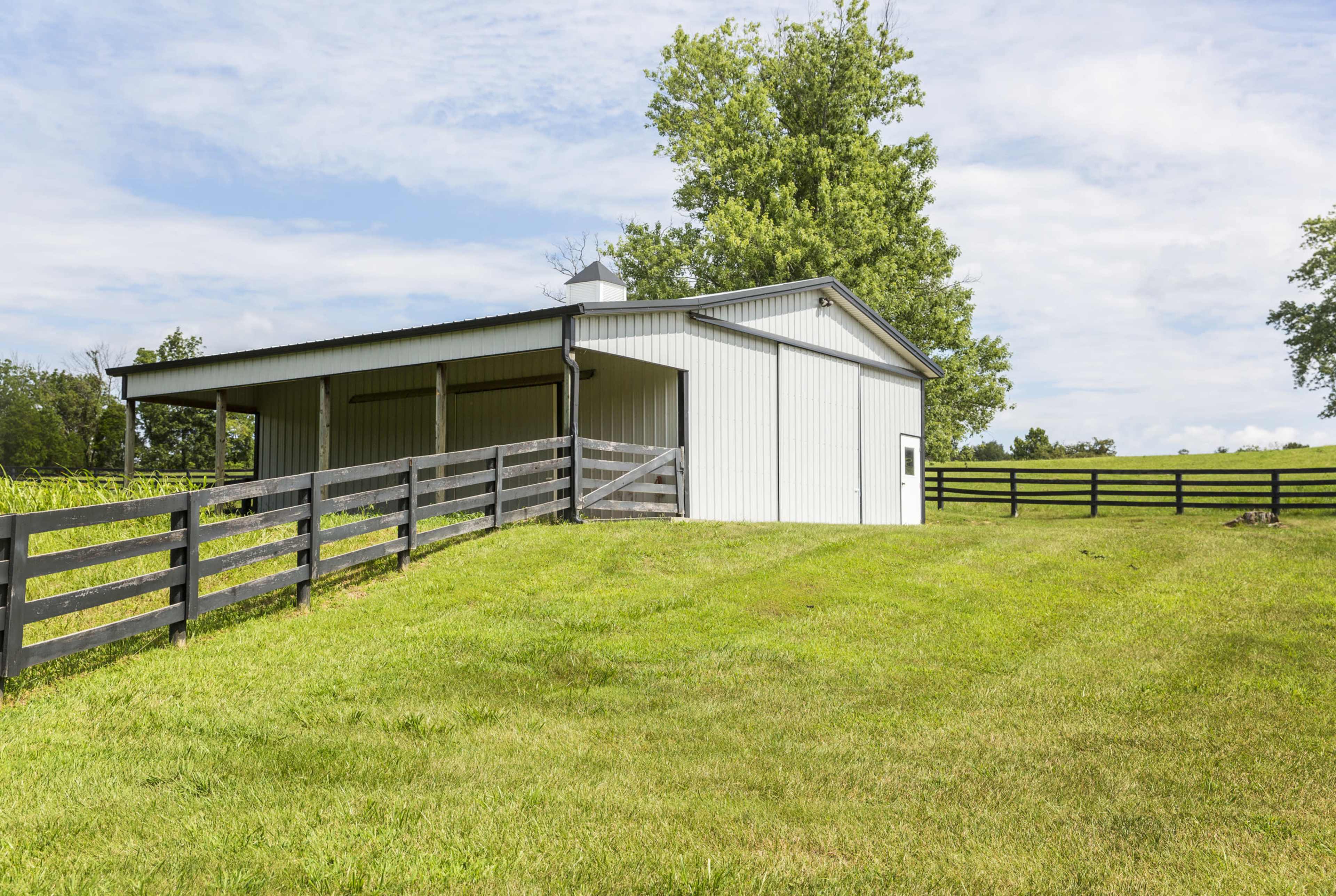 A metal barn situated on a grassy slope, surrounded by a black wooden fence and tall trees in the background.
