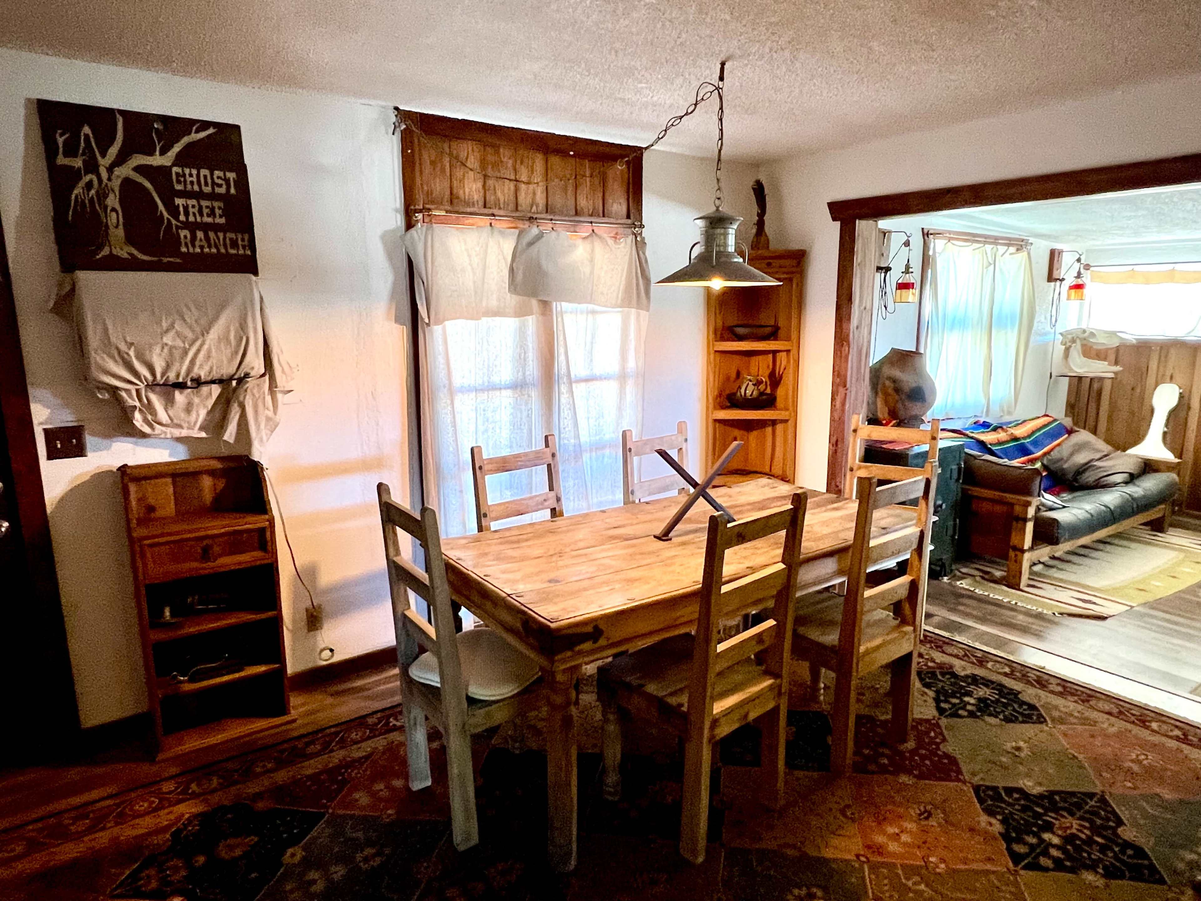 A rustic dining area features a wooden table surrounded by chairs, with a hanging light overhead and a view into a brightly lit adjacent room.