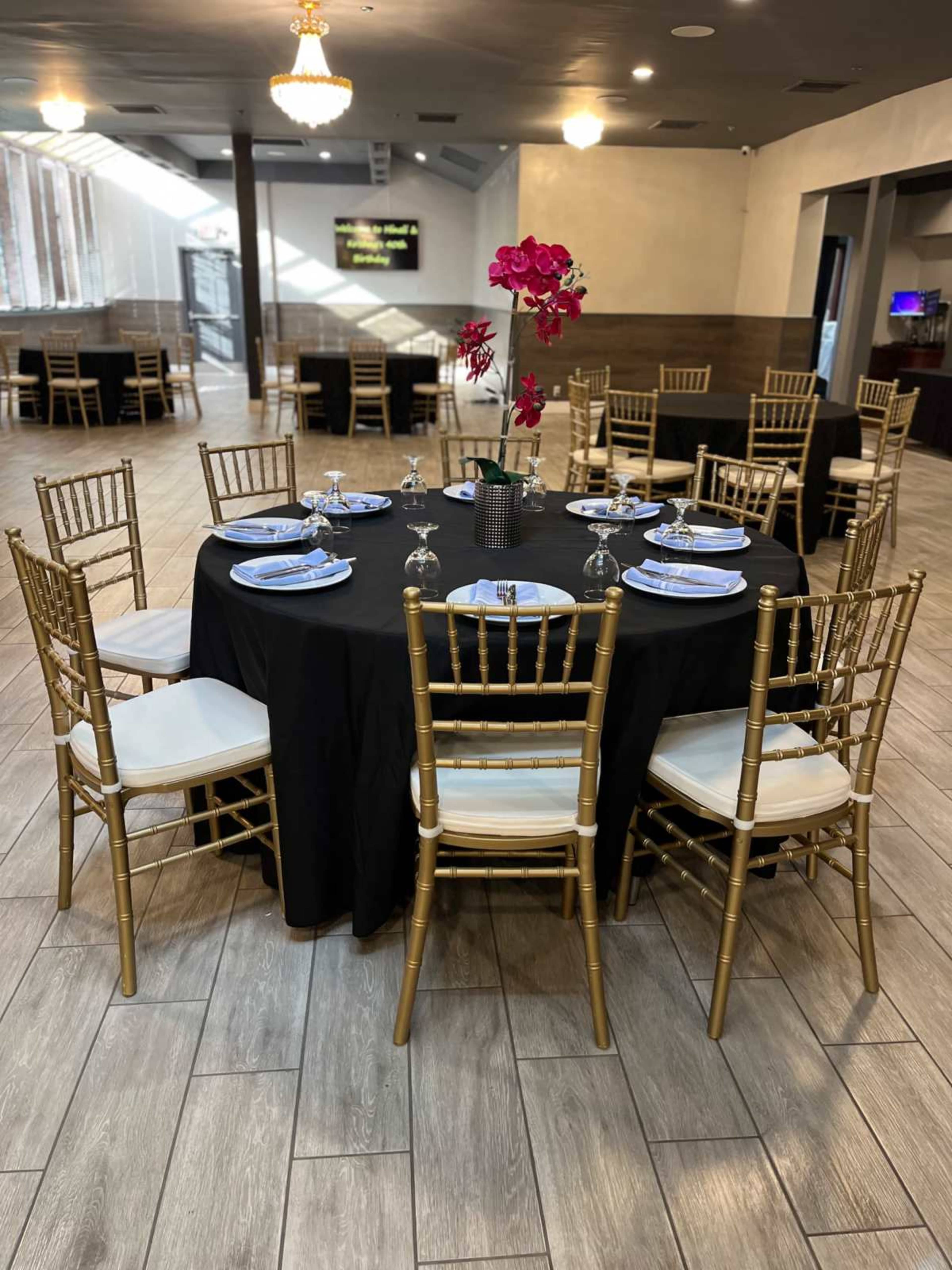 A round table is set with blue plates and silverware, adorned with a flower centerpiece, in a spacious dining area featuring gold chairs and a dark tablecloth.