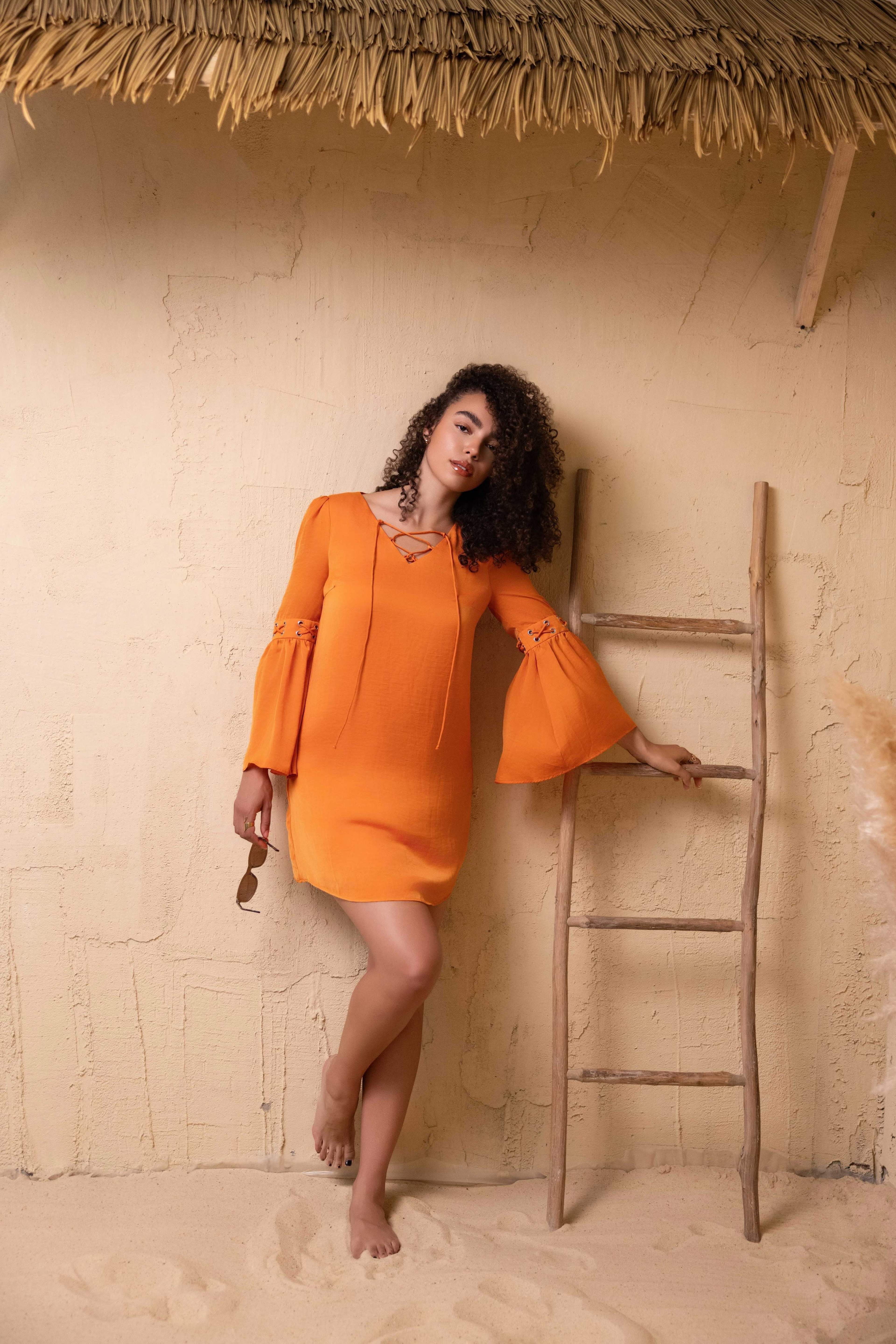 A woman in an orange dress with bell sleeves poses next to a wooden ladder against a textured wall in a beach setting.