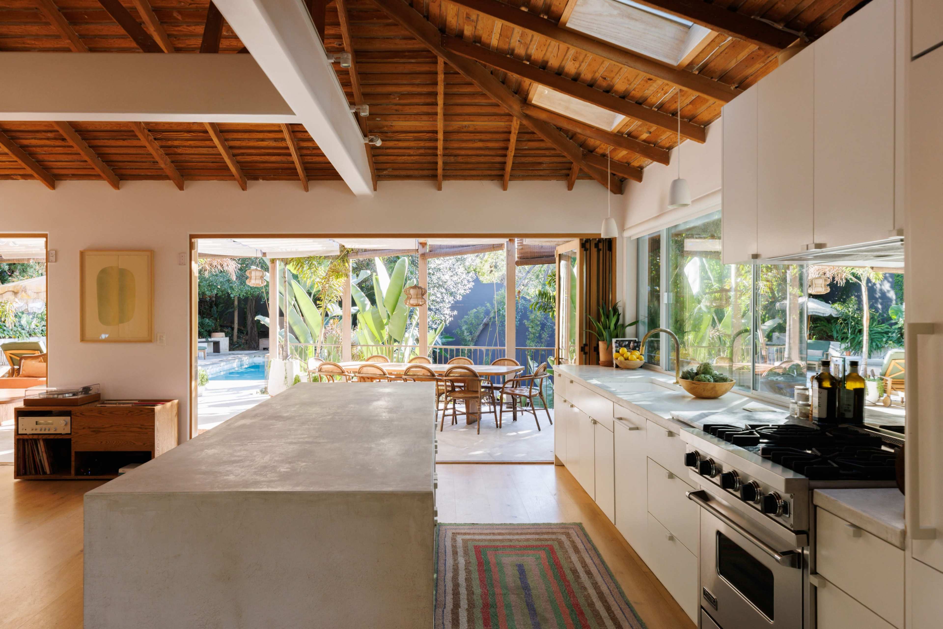 The image shows a modern kitchen with a concrete island, white cabinetry, and large windows that open to a patio area with outdoor seating and a pool.
