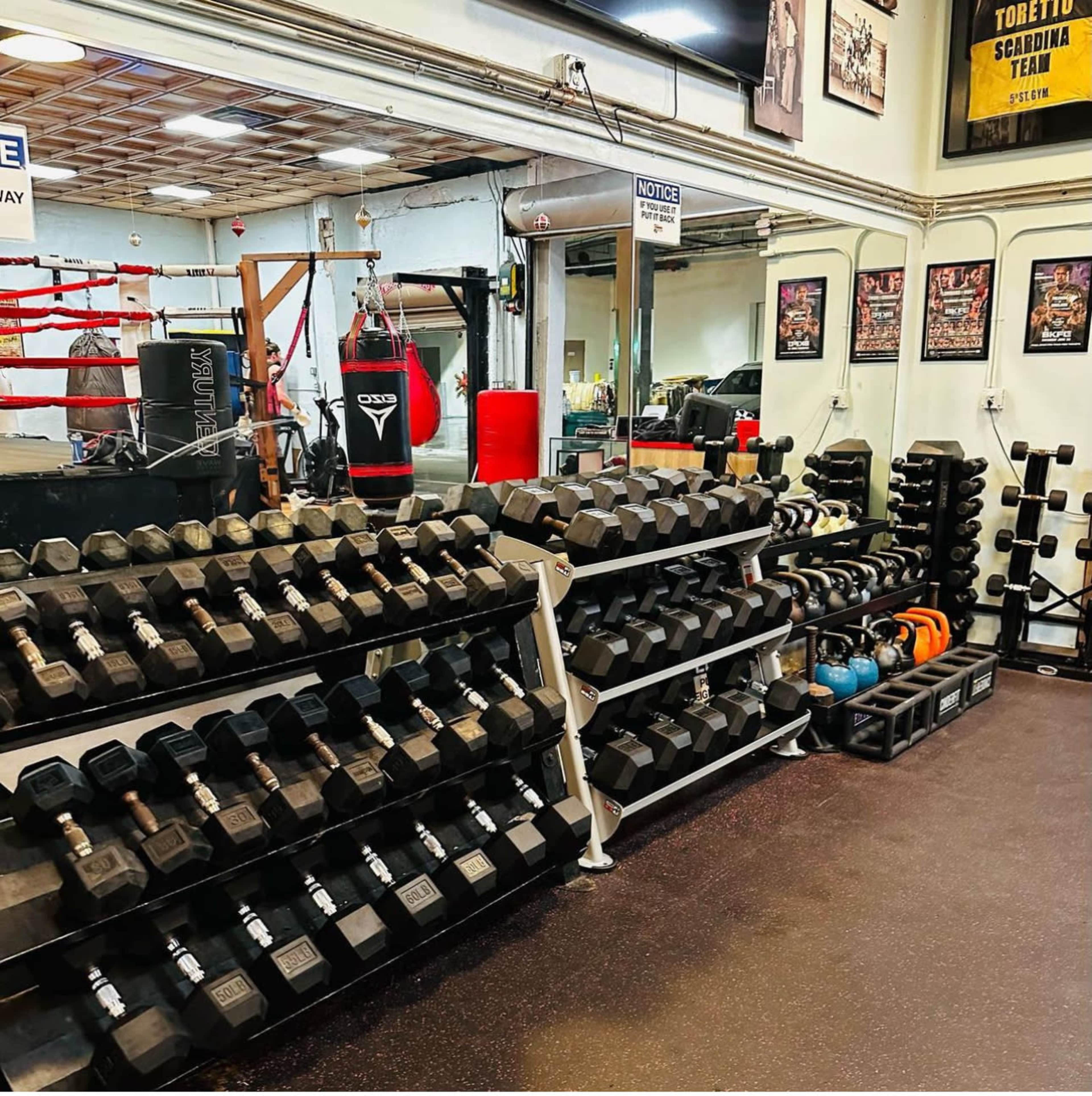 A gym interior with a rack of dumbbells, kettlebells, and a boxing setup including a heavy bag and a ring.
